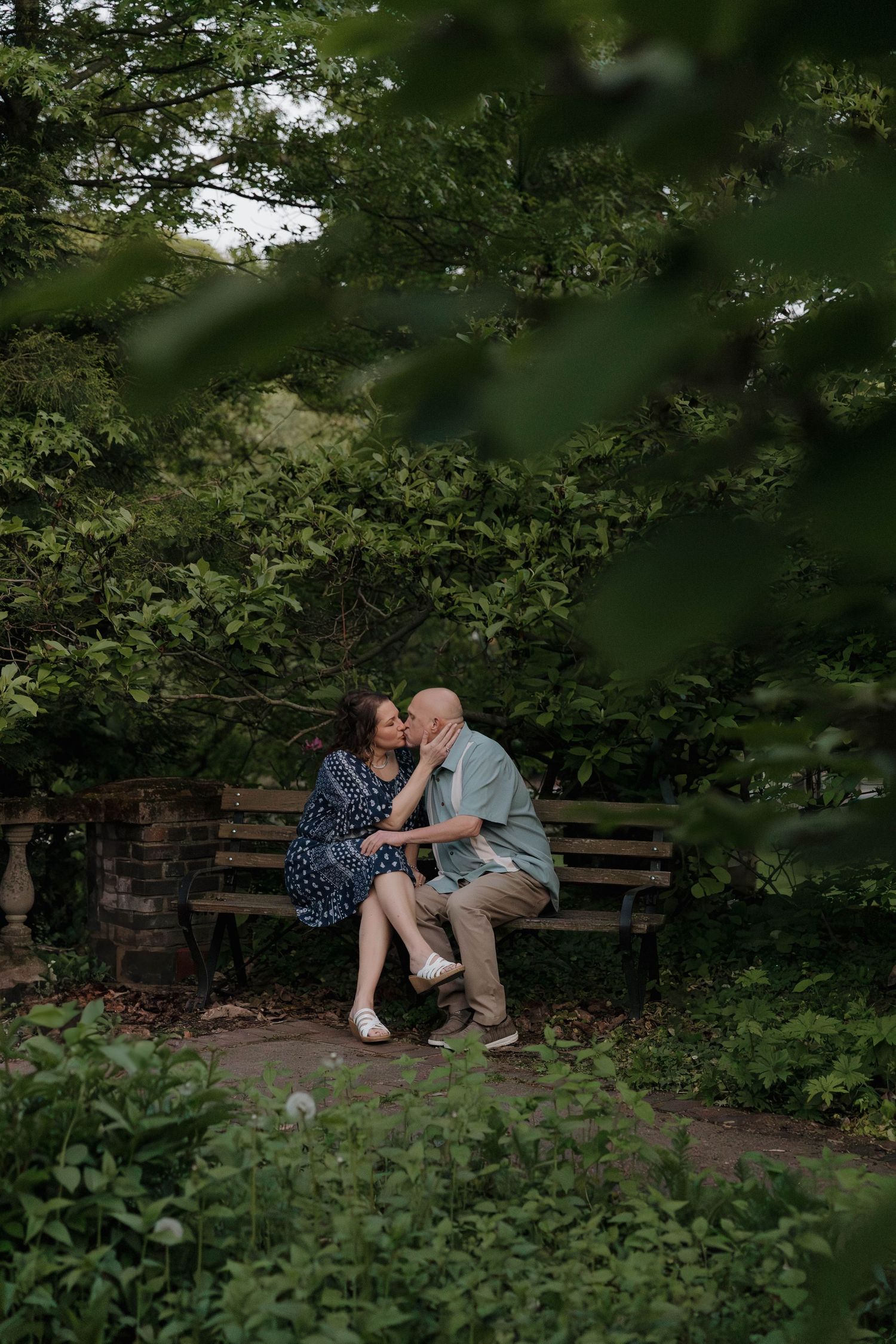 A couple shares an intimate moment on a rustic wooden bench surrounded by lush green foliage in a garden setting.