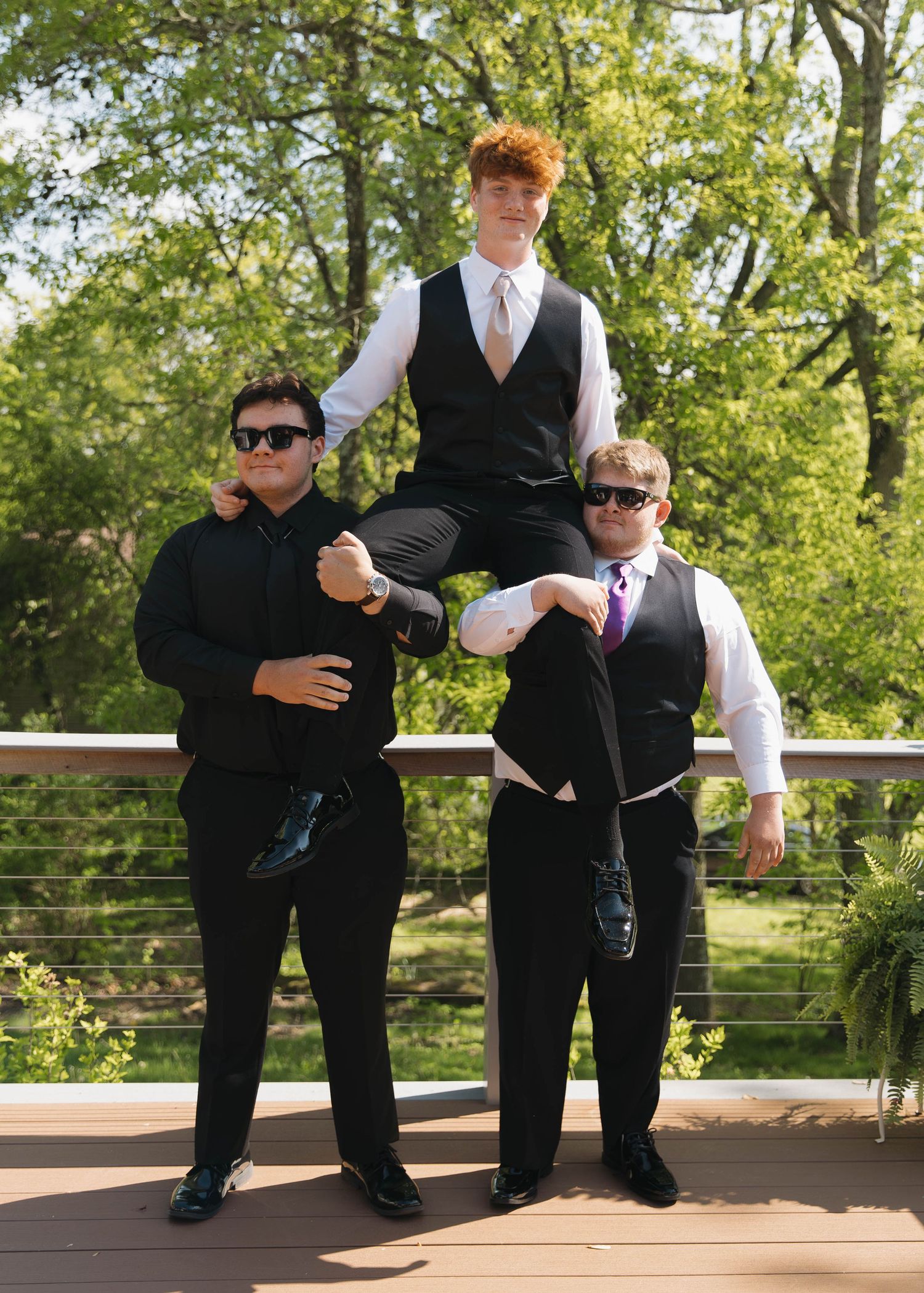 Three formally dressed people in black suits and ties pose together on a wooden deck with green trees in the background.