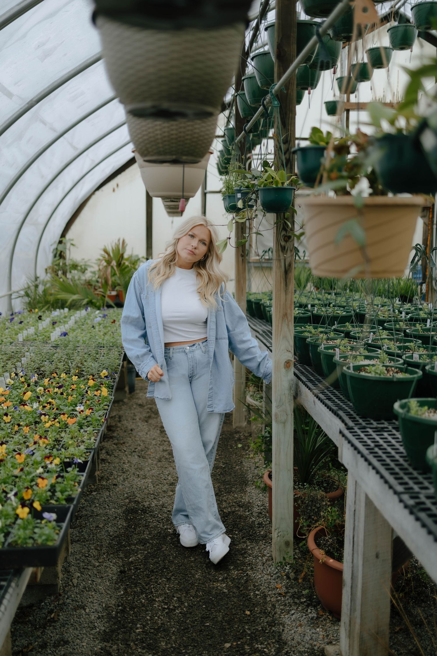 A greenhouse interior with potted plants and flowers on shelves, featuring light denim clothing and white sneakers.