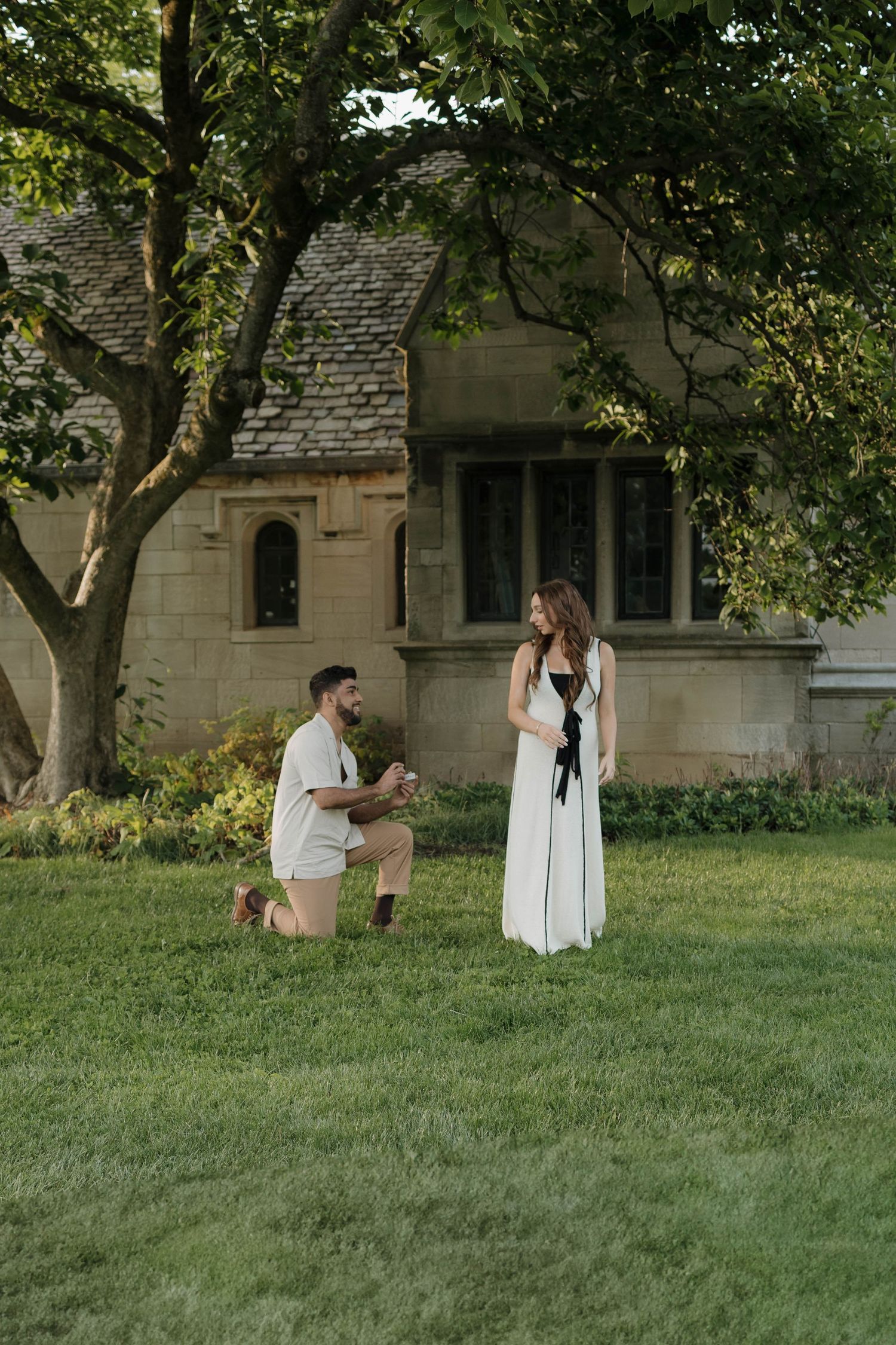 A romantic proposal moment on a lush green lawn in front of a stone church building during summer.