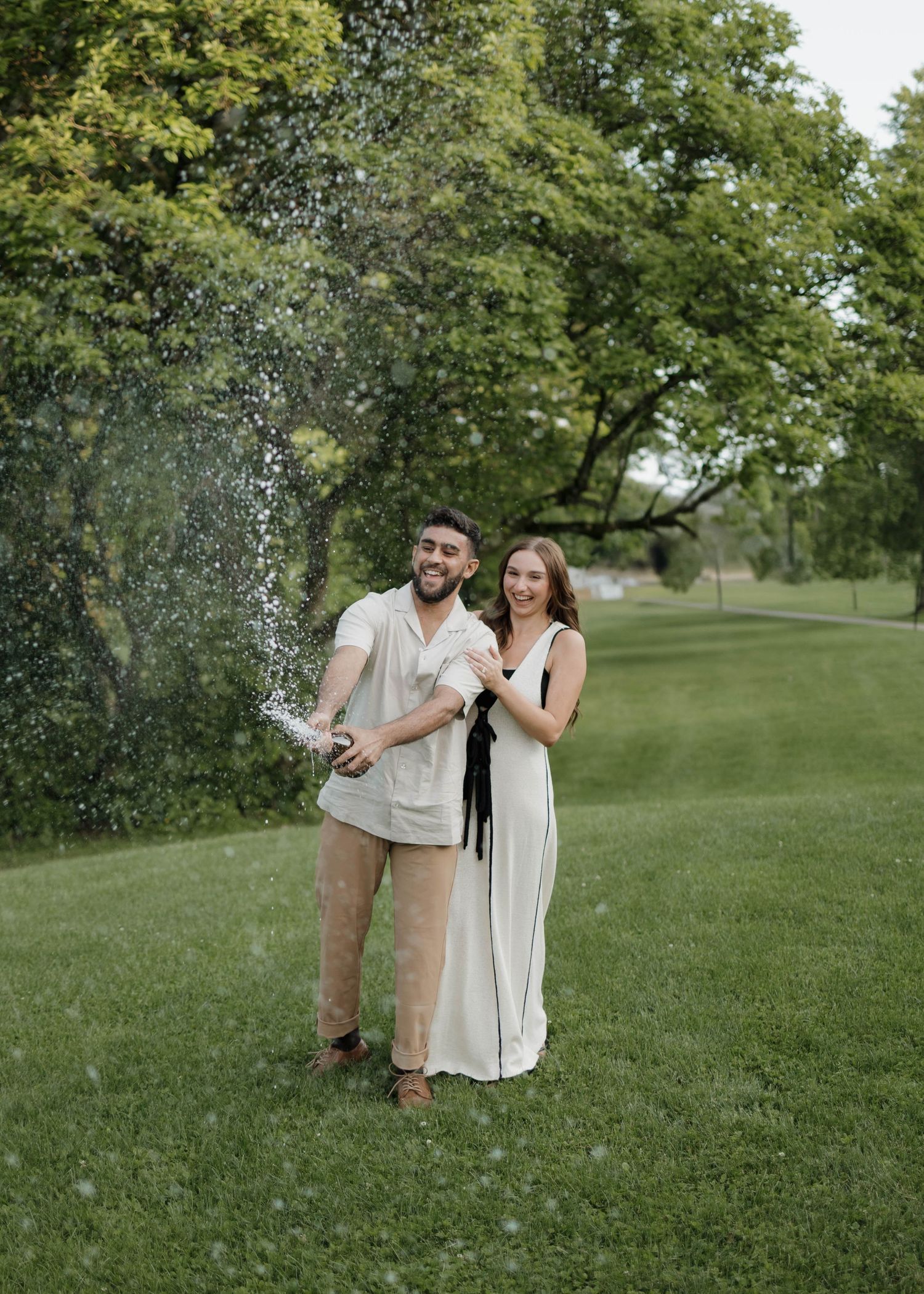 Couple celebrates with champagne spray in a lush green garden setting wearing coordinated neutral-toned outfits.
