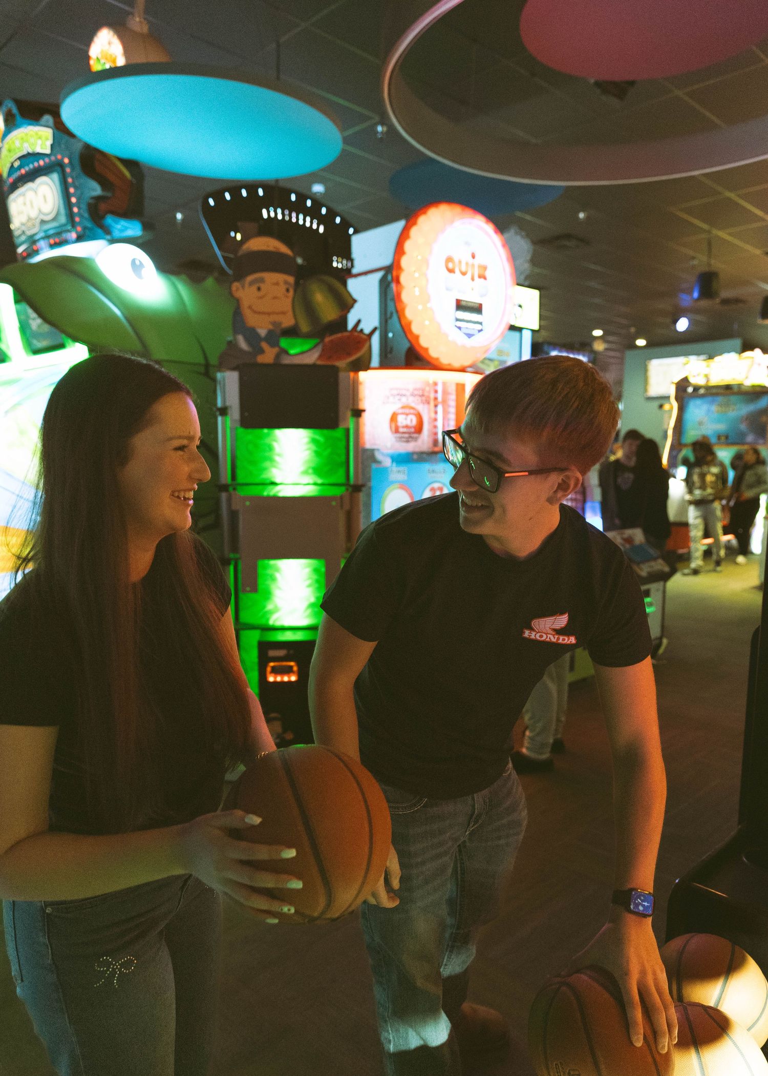 Two employees share a laugh while working at an arcade with illuminated gaming machines in the background.