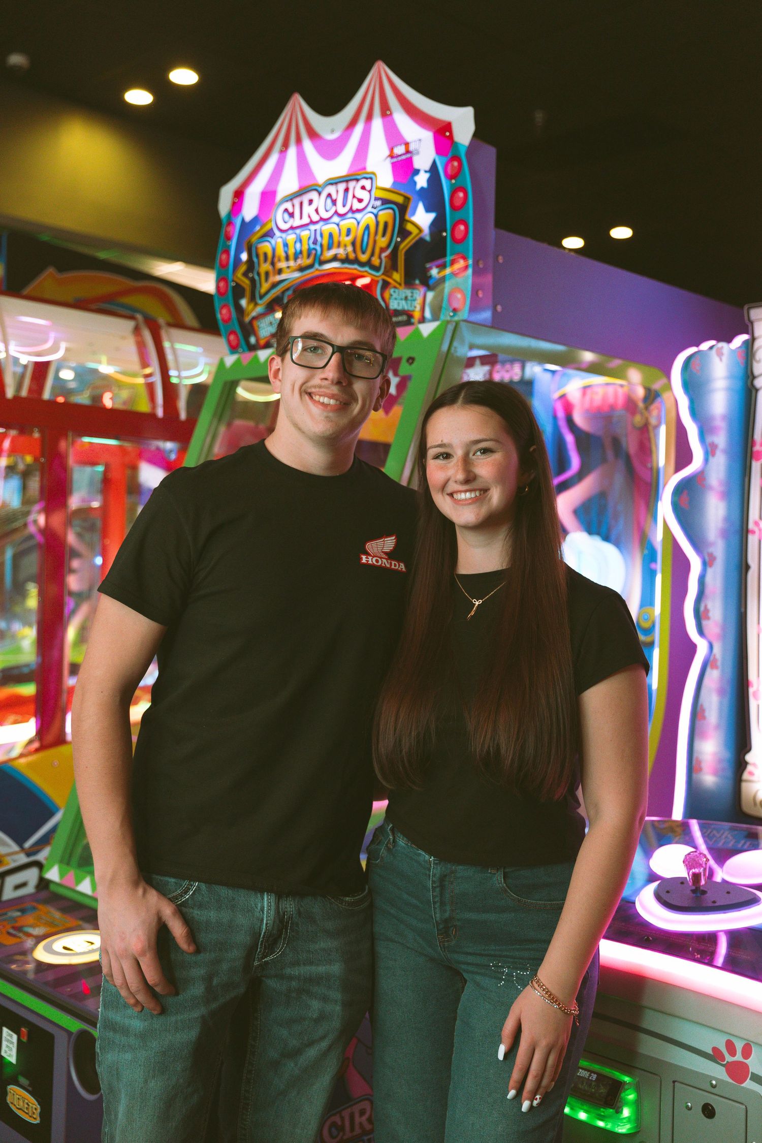Two staff members in black t-shirts stand smiling in front of colorful arcade game machines.
