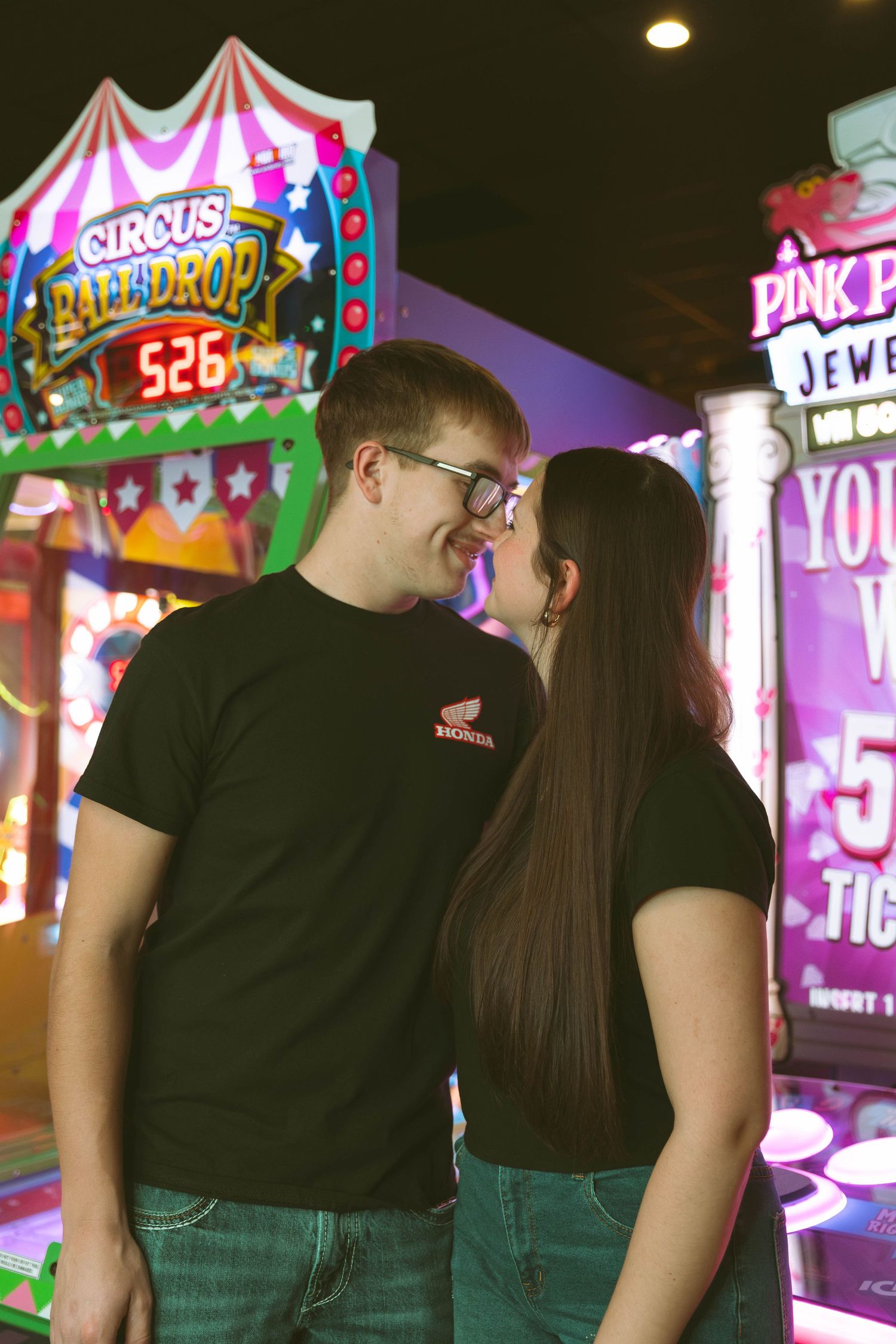 A couple in matching black t-shirts gazes at each other in a colorful arcade with bright neon game signs glowing behind them.