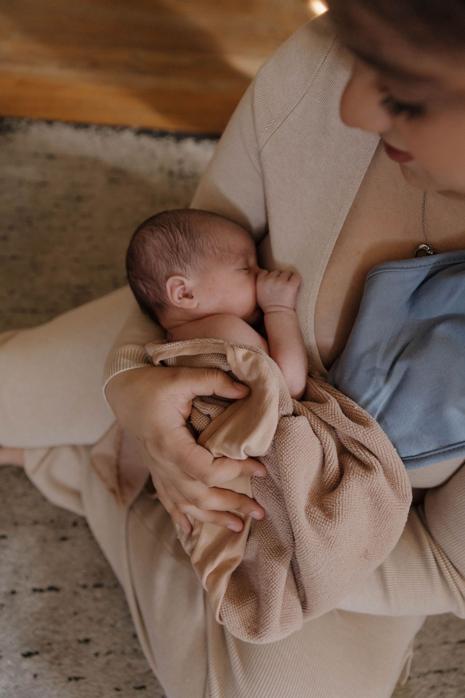 A newborn baby wrapped in beige knit blanket nurses while being cradled in gentle embrace.