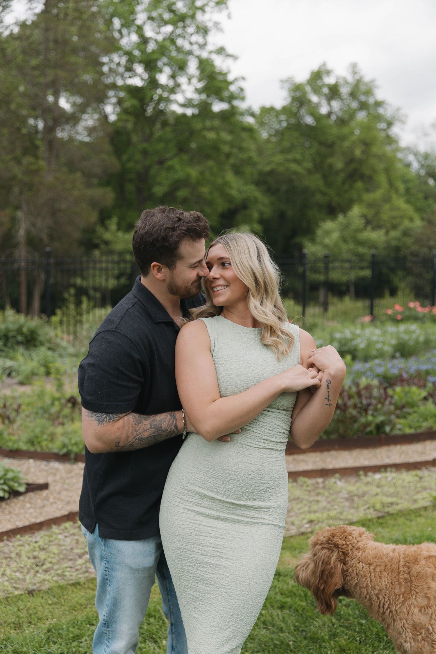 A romantic couple embraces in a garden setting with a brown dog visible in the background.
