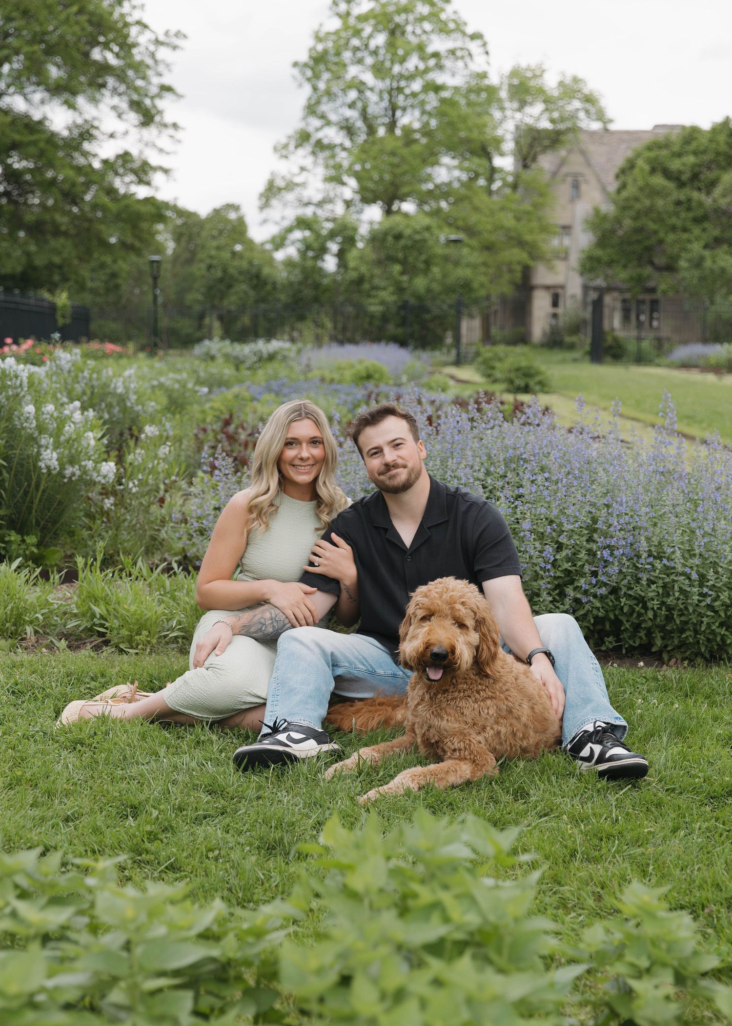 Goldendoodle dog laying on grass next to couple in garden with purple flowers and green trees in background.
