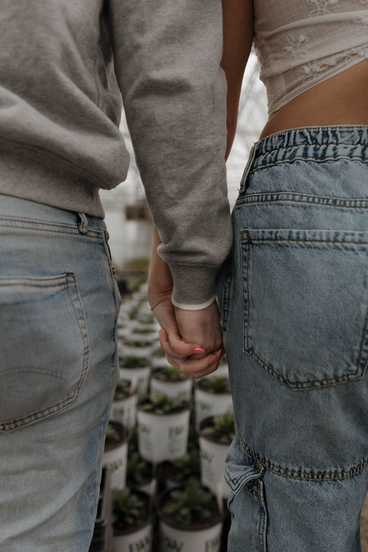 A couple holding hands while wearing casual jeans and a gray sweatshirt, standing near plant containers.