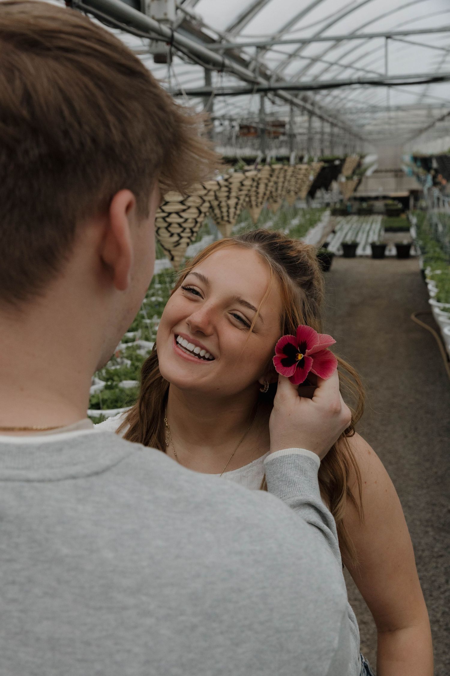 A couple shares a tender moment in a greenhouse as someone places a vibrant red flower in their companion's hair.