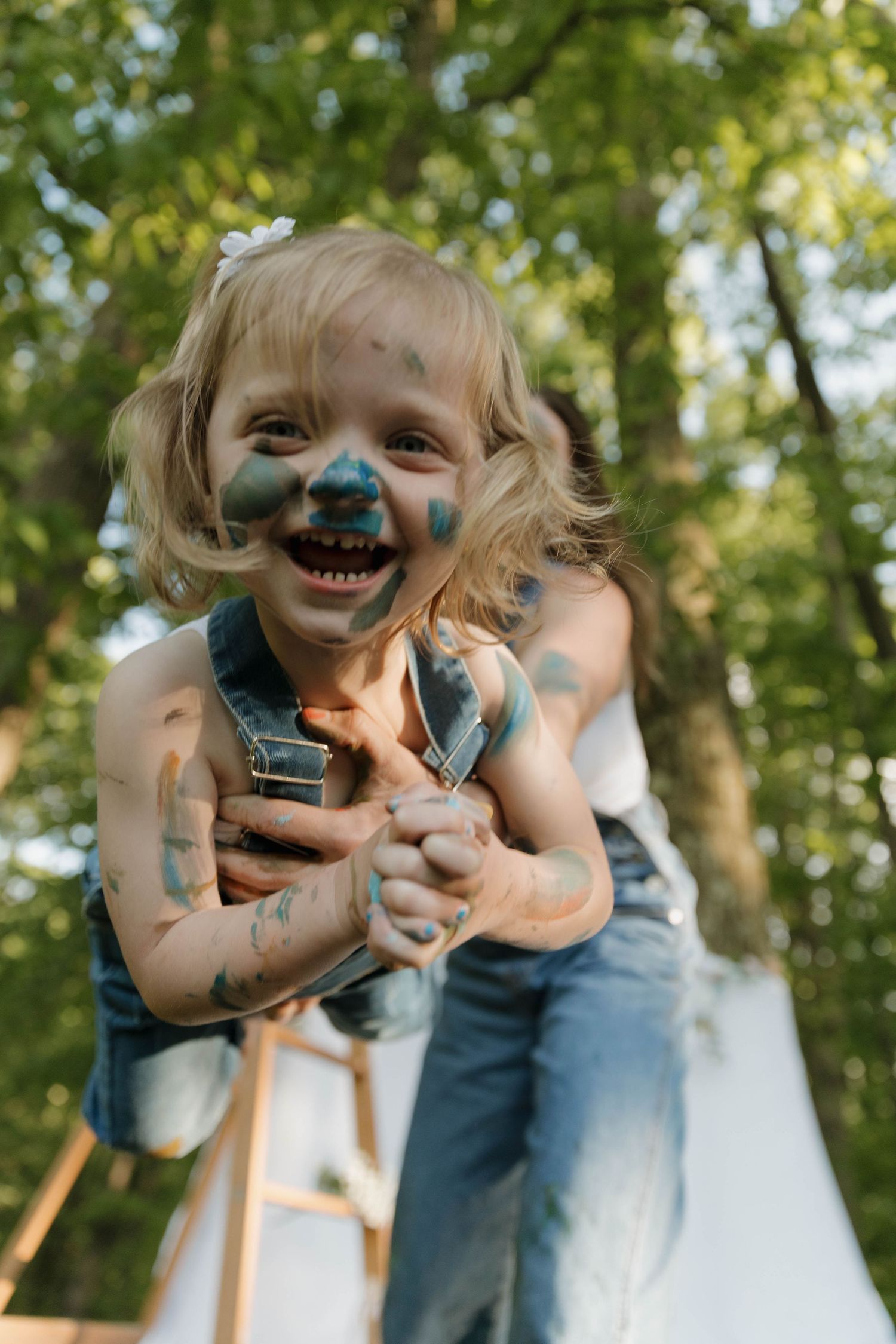 Joyful child playing outdoors with face paint, wearing overalls while being lifted up against a leafy green background.