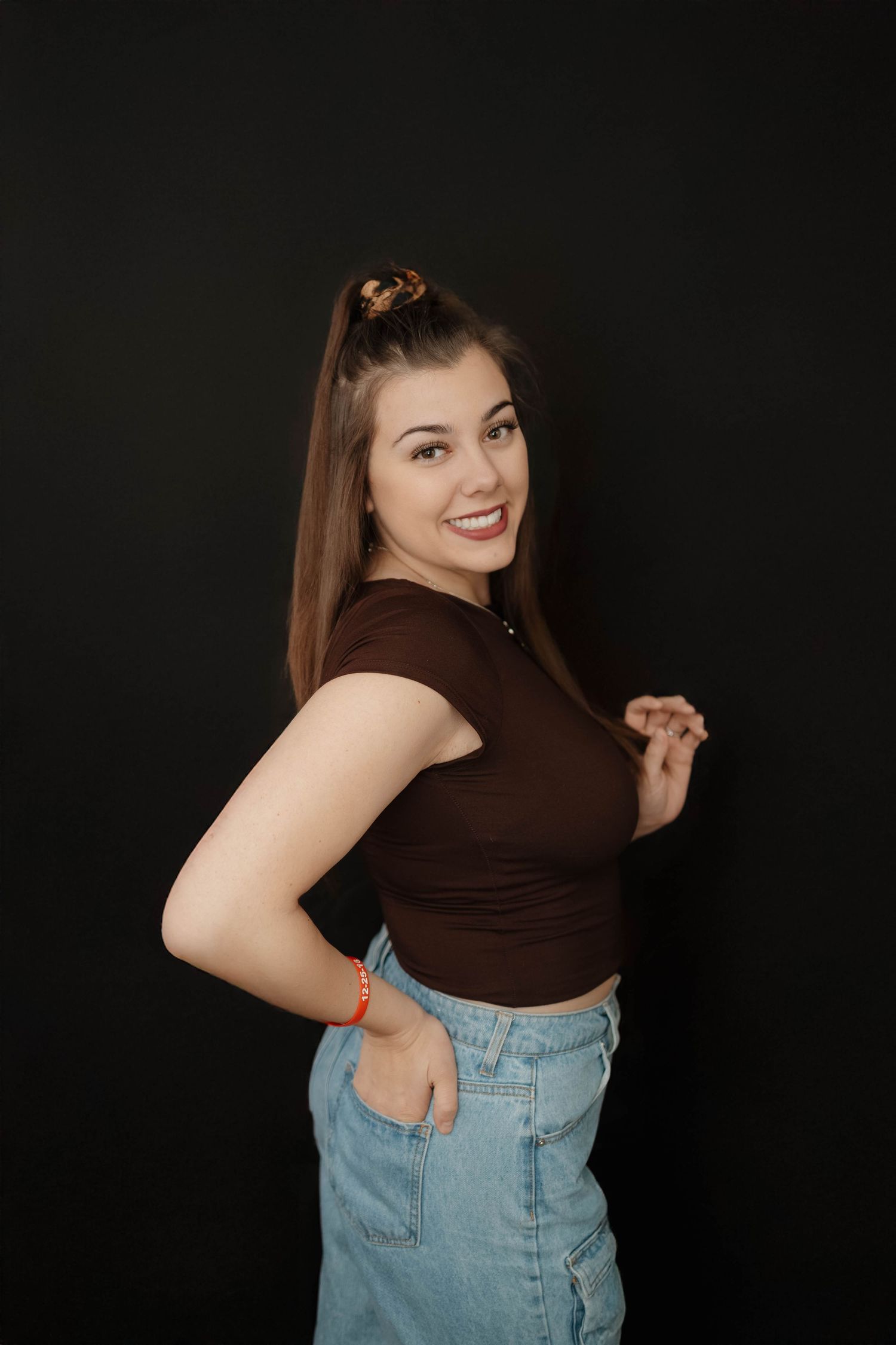 A smiling person in a brown crop top and light denim jeans poses against a dark background.