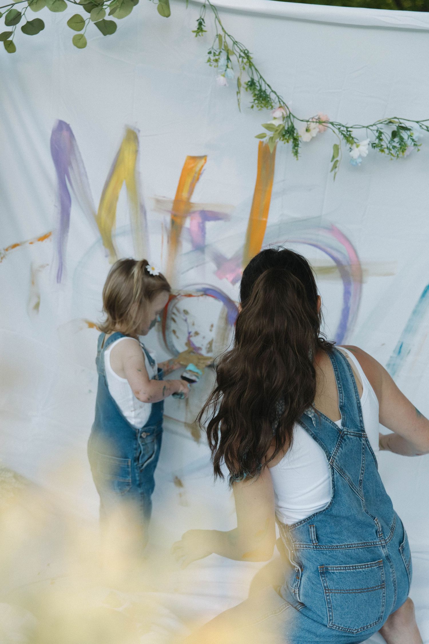 Two people in denim overalls paint together on a white wall outdoors with green foliage visible above.