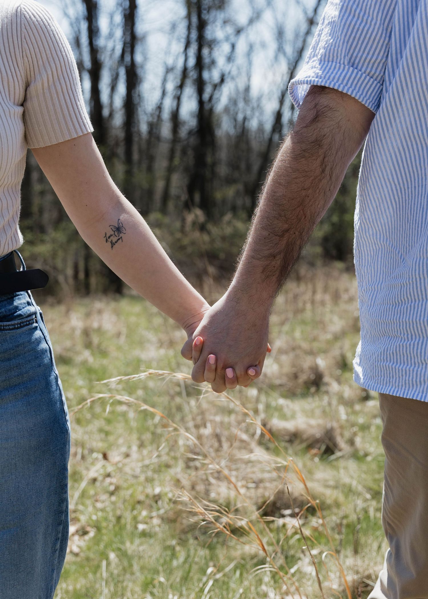 Two people holding hands intimately in a sunlit field with bare trees in the background.