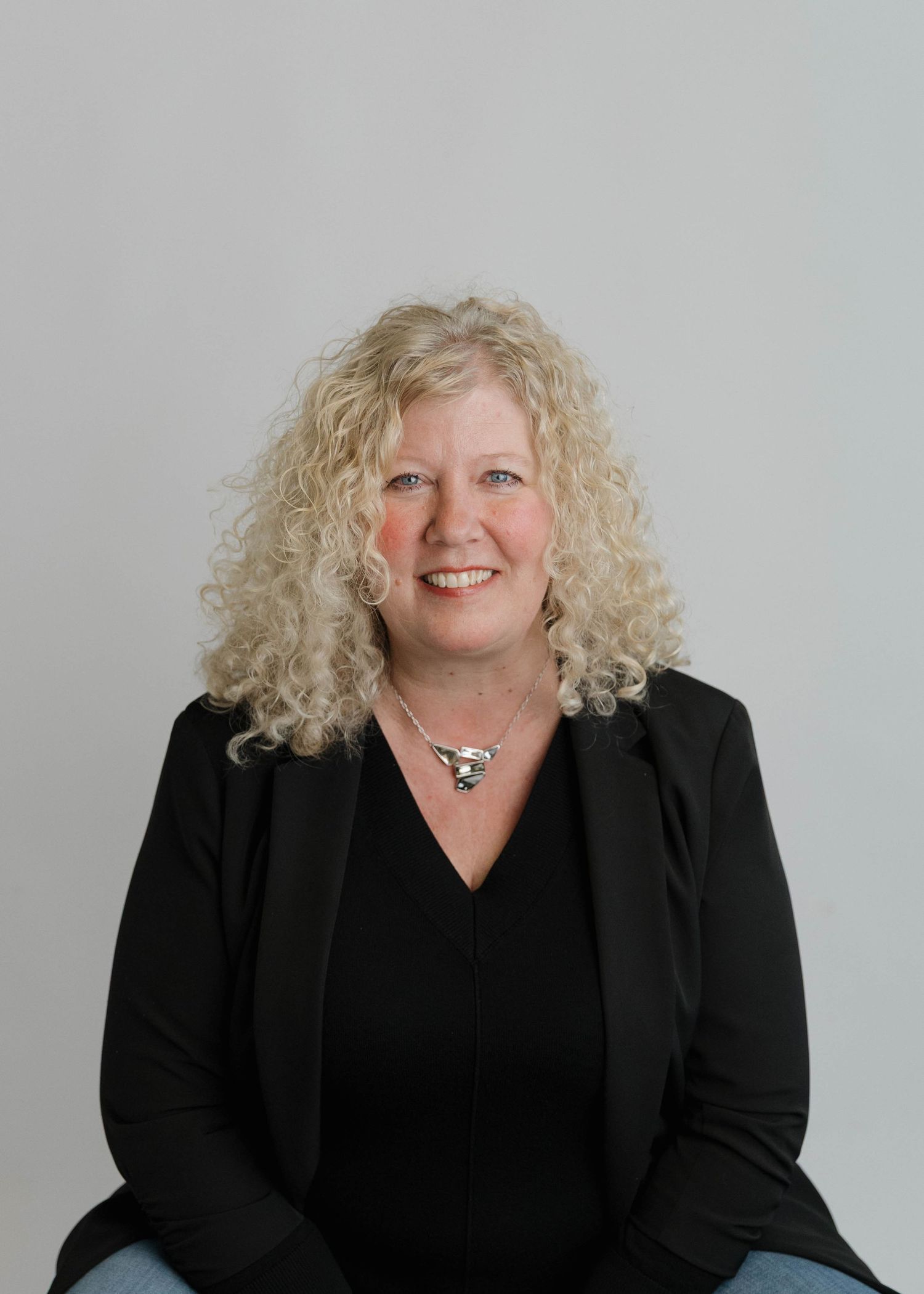 Professional headshot of someone with curly blonde hair wearing a black blazer and top against a light gray background.