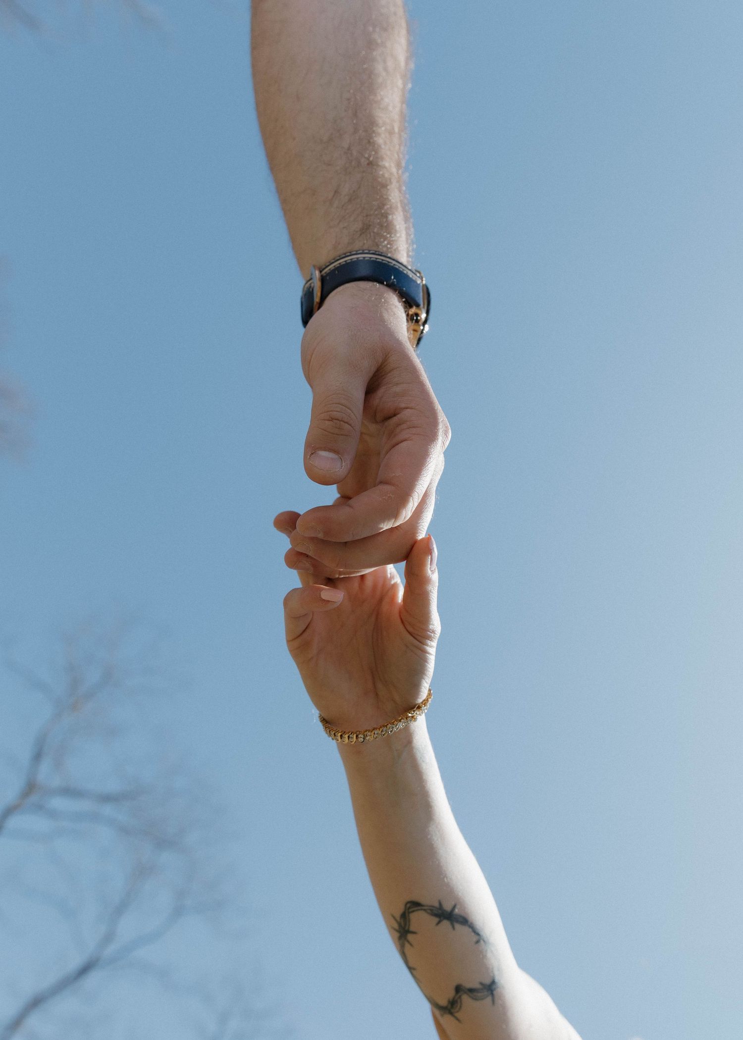 Two hands reaching and clasping together against a blue sky, with one arm showing a small tattoo.