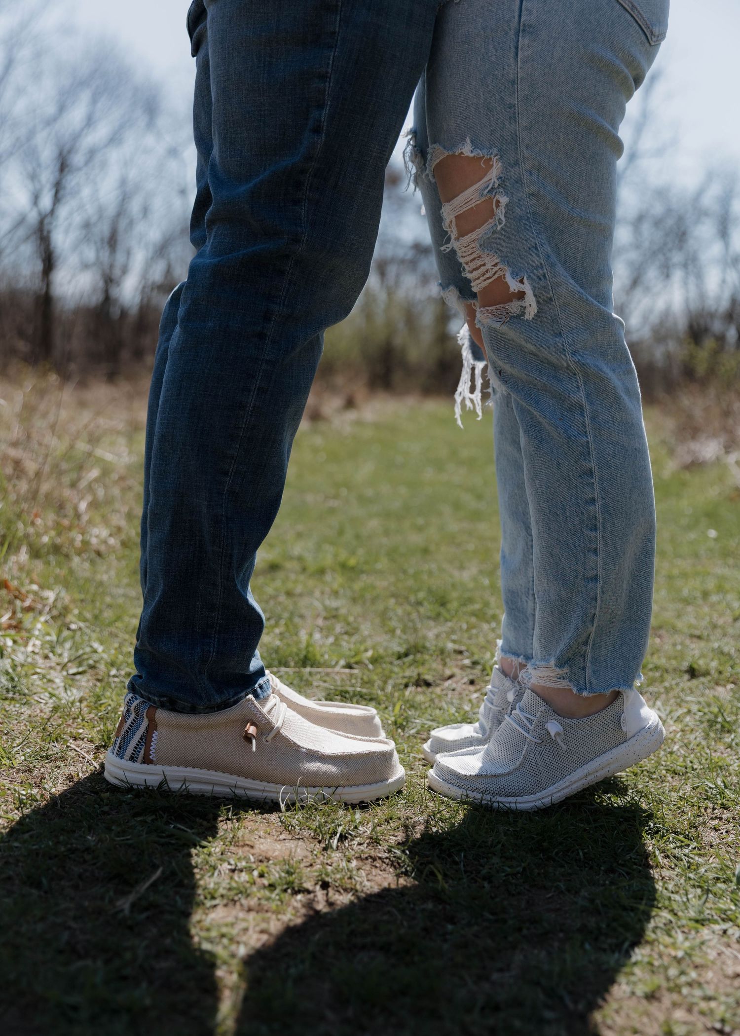 Two people wearing contrasting shoes - brown loafers and white sneakers - stand close together in grass on a sunny day.