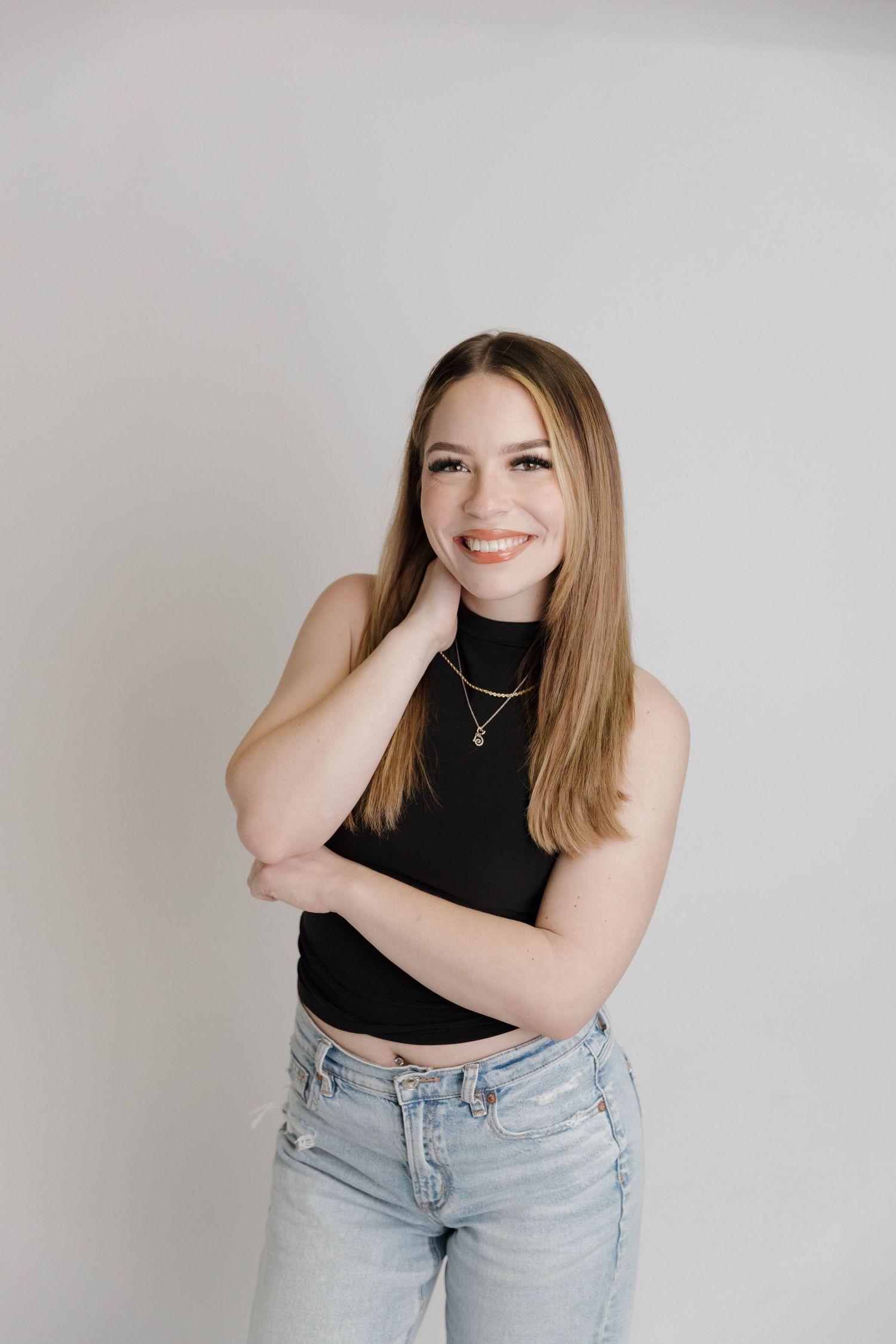 A person wearing a black crop top and jeans poses casually with hand on neck against white backdrop.