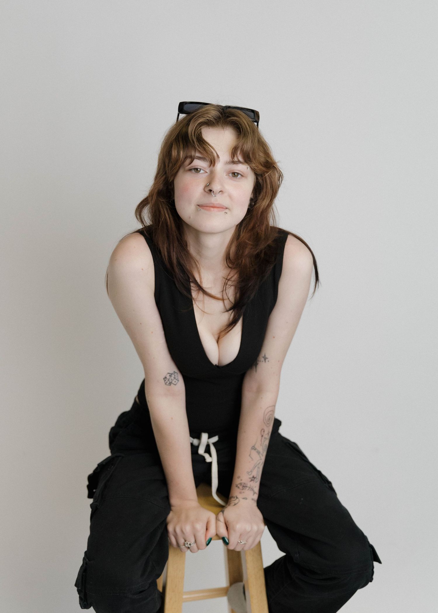 A person in a black outfit sits casually on a wooden stool against a light gray background.