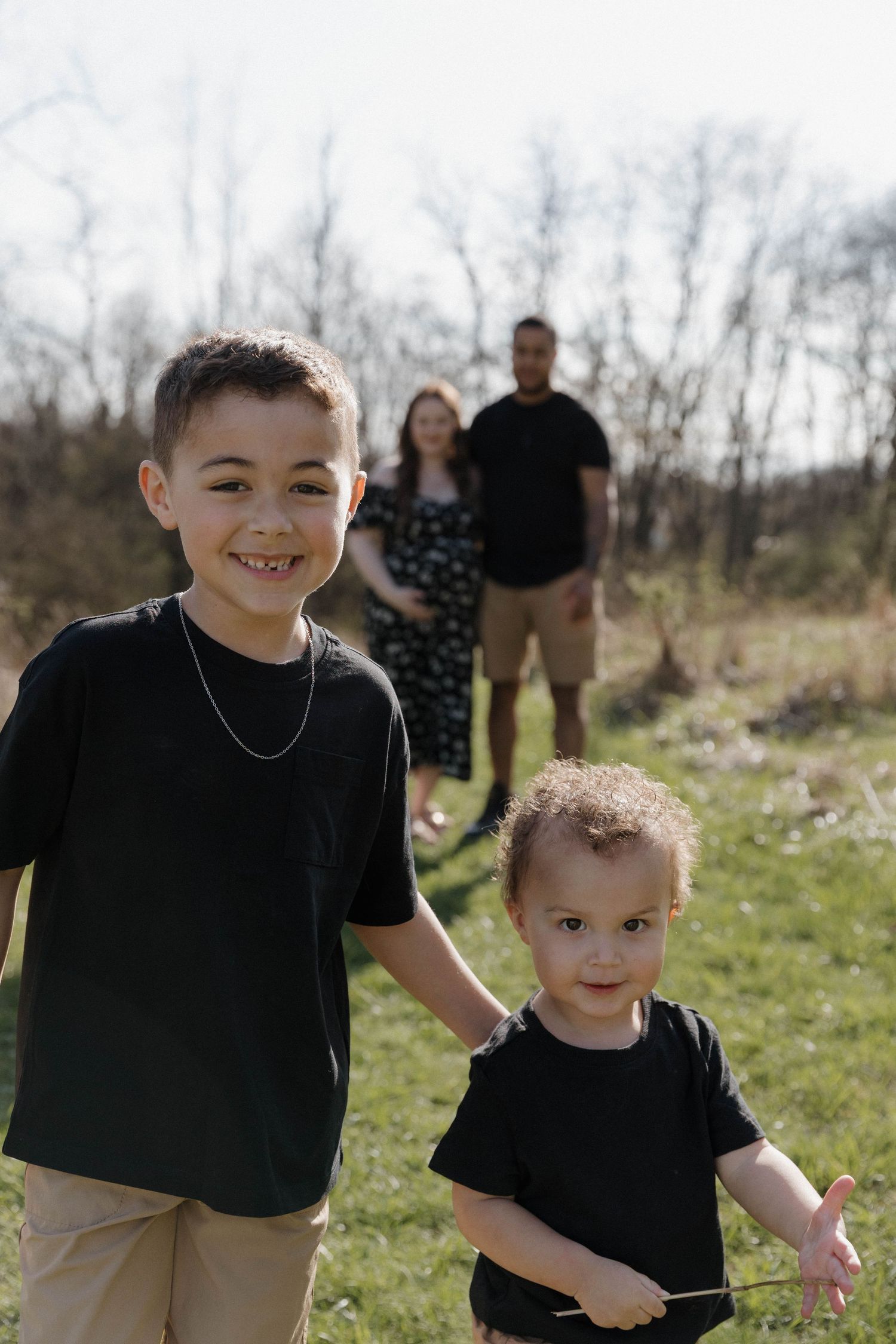 Two children in black shirts stand in a sunny field with family members visible in the background.