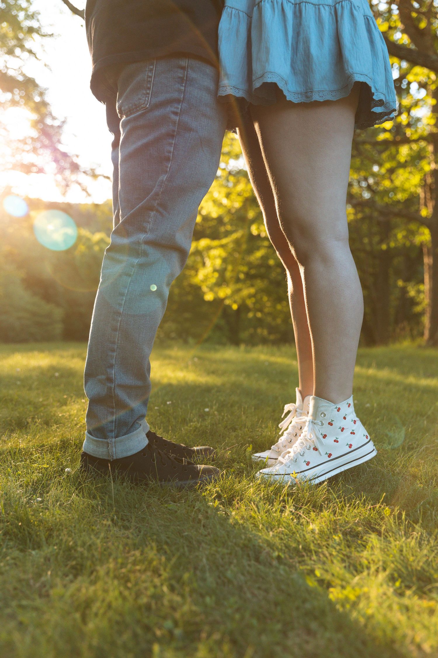 Two pairs of legs standing close together in sunlit grass, one wearing jeans and dark shoes, the other in a blue skirt and white sneakers.