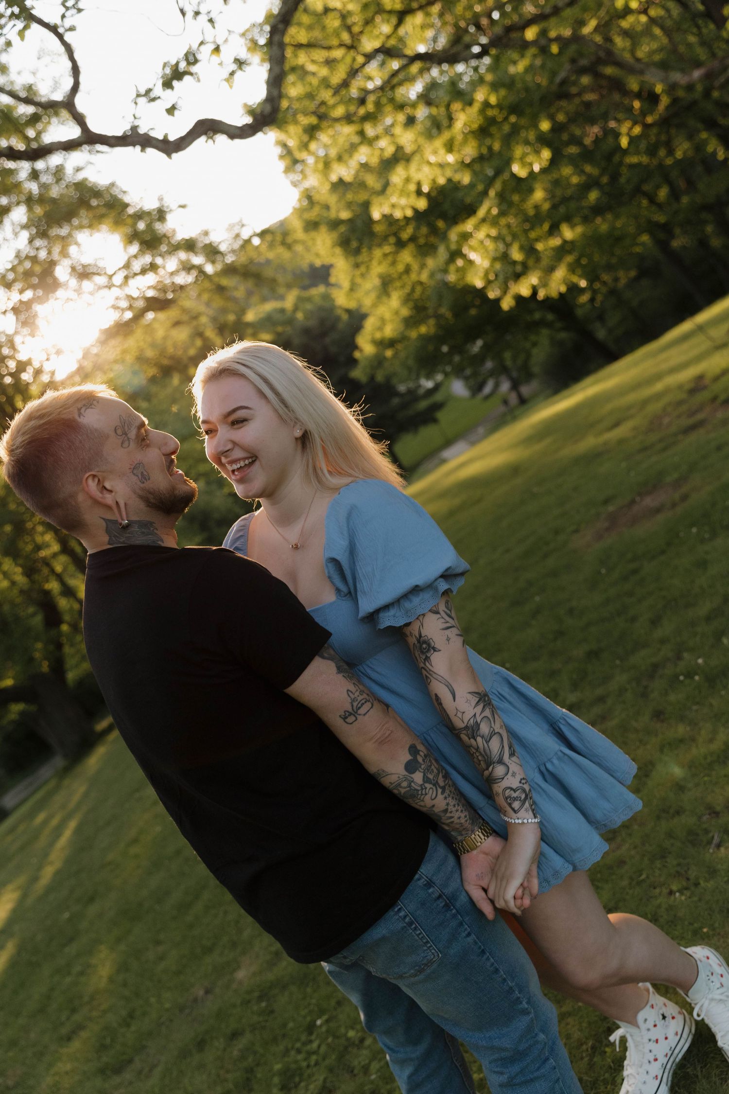 A couple shares a tender moment in a sunny park setting during golden hour, with lush greenery in the background.