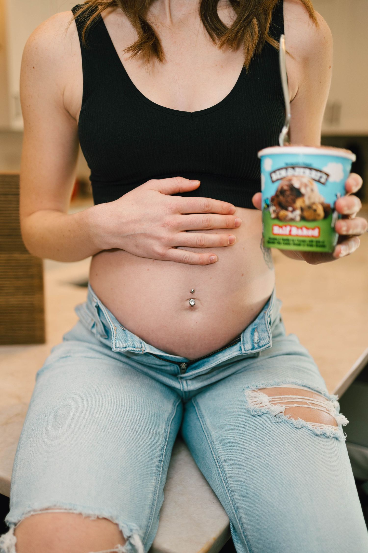 Person wearing black tank top and ripped jeans holds a Ben & Jerry's ice cream container while showing off their navel piercing.