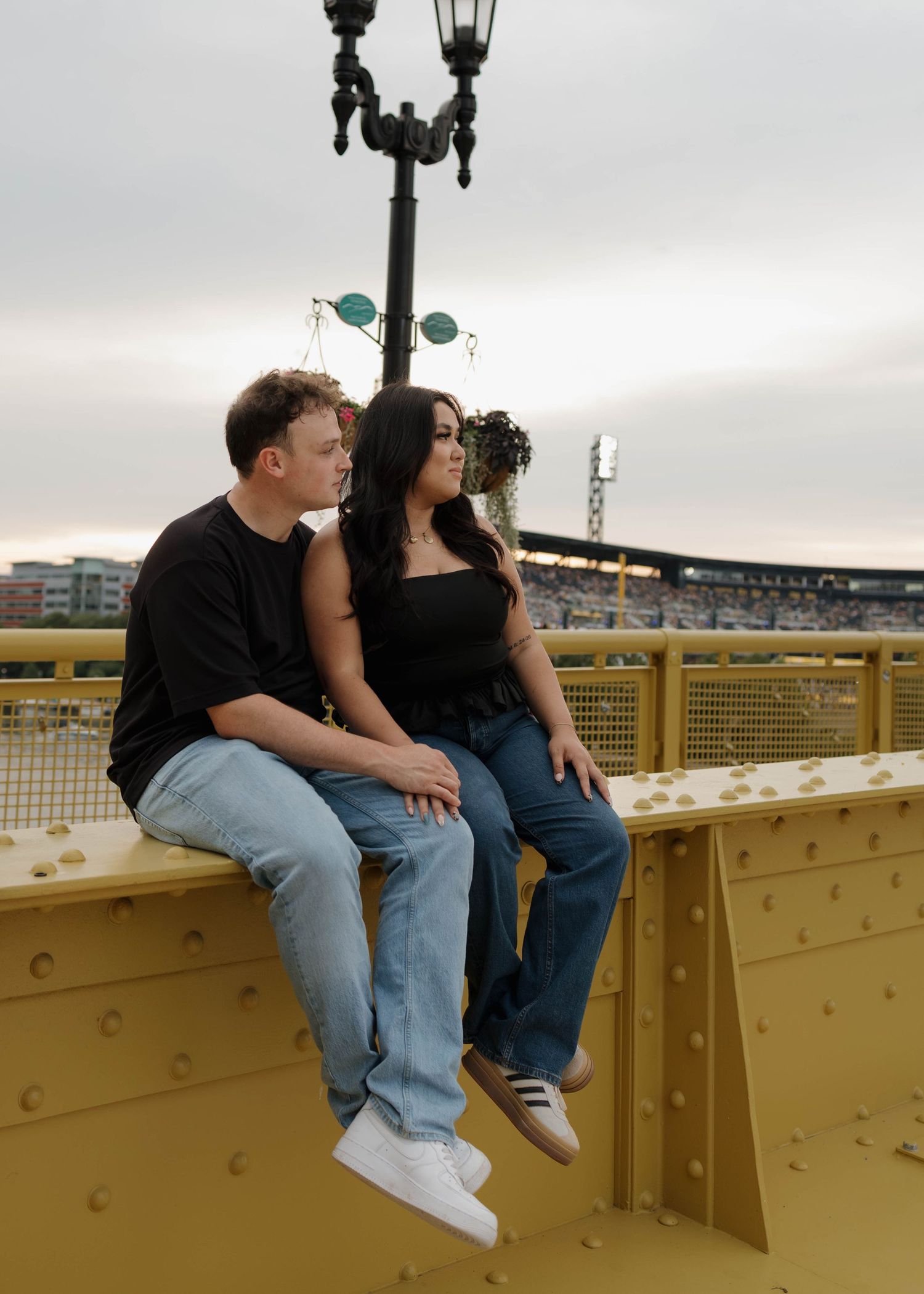A couple sits together on yellow bridge railing at dusk with decorative lamp post and stadium lights in background.