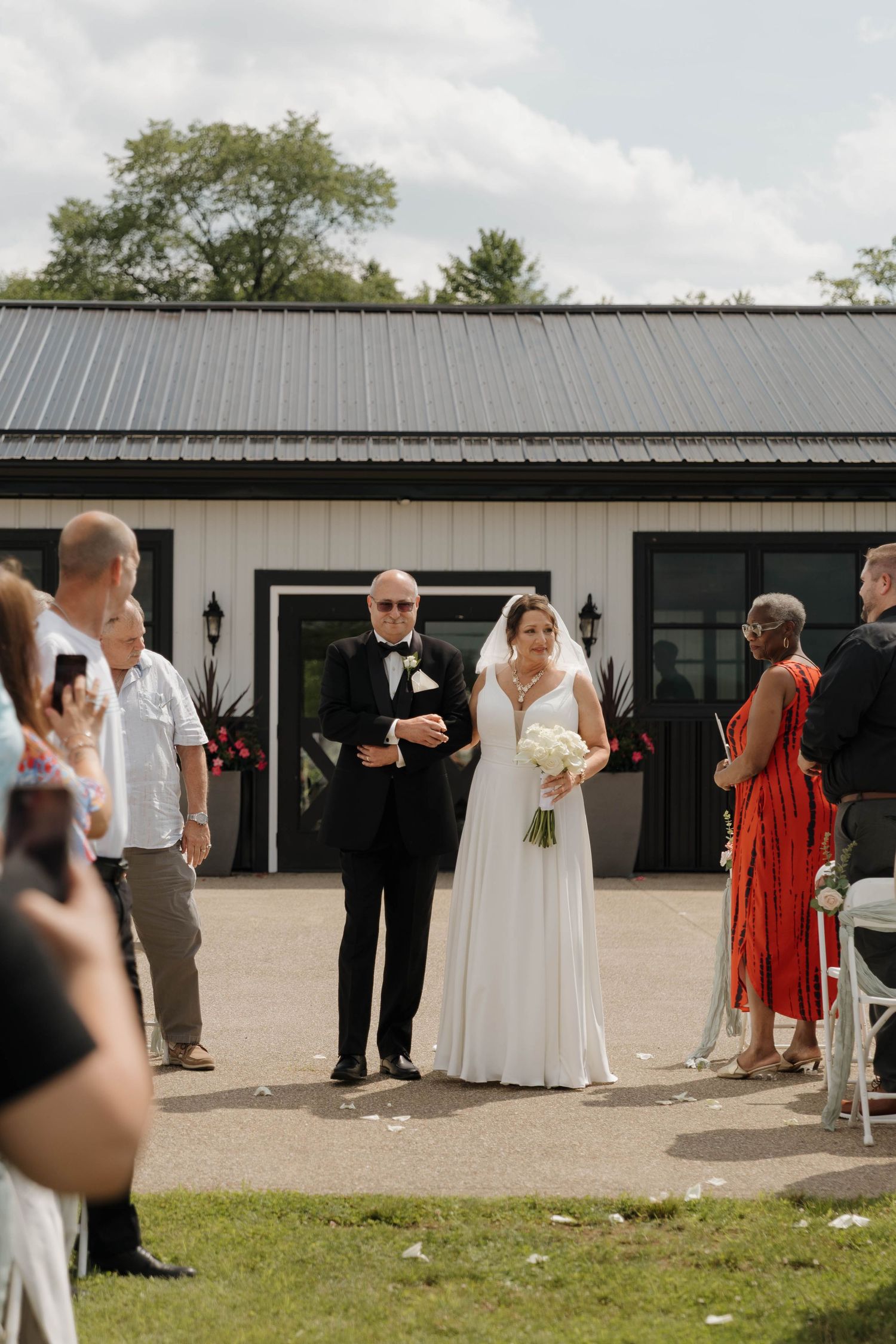 Wedding ceremony guests gather outside a modern venue with metal roofing and white exterior on a sunny day.