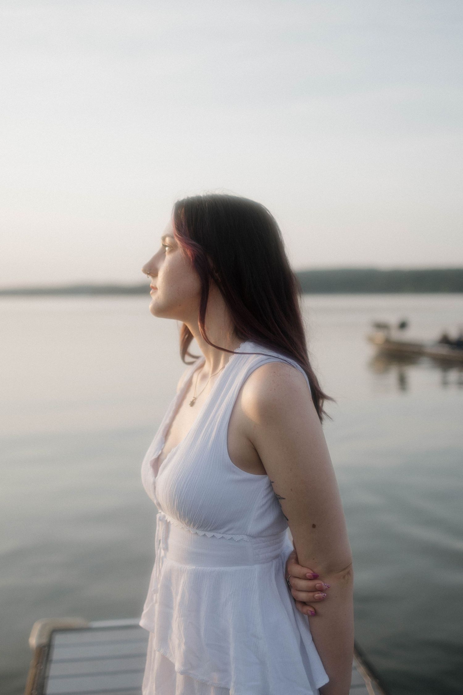 Person in a white sundress stands on a dock by a serene lake at sunset while boats float in the distance.