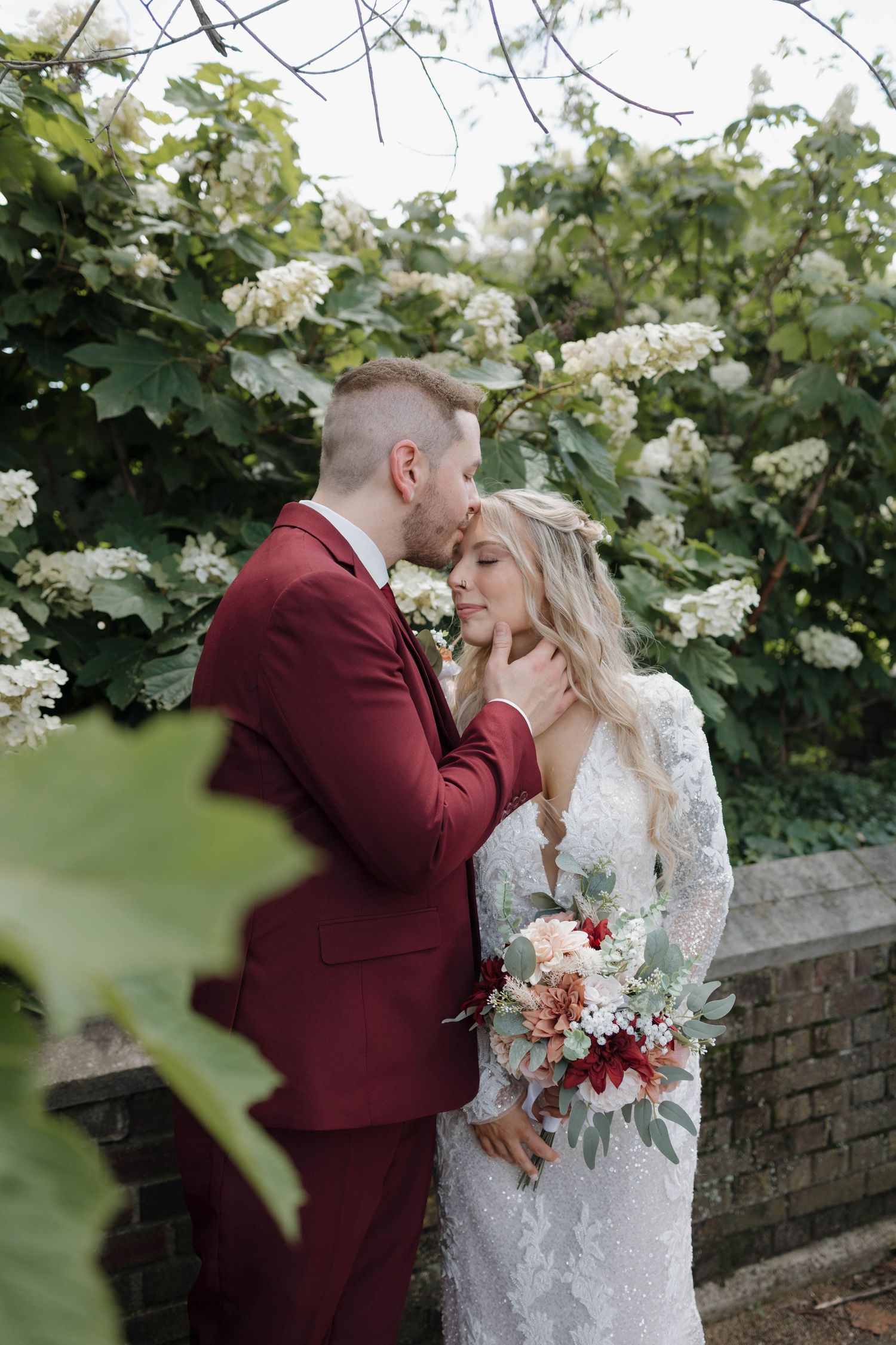 A bride in white lace and groom in burgundy suit share an intimate moment surrounded by blooming floral vines.