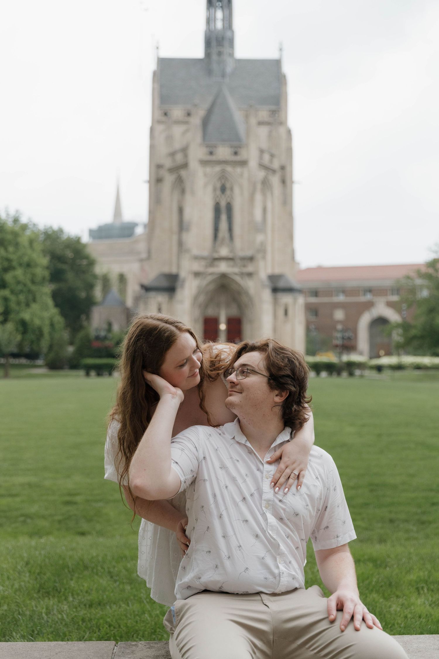 A couple shares tender moments on a green lawn with a Gothic cathedral building visible in the background.