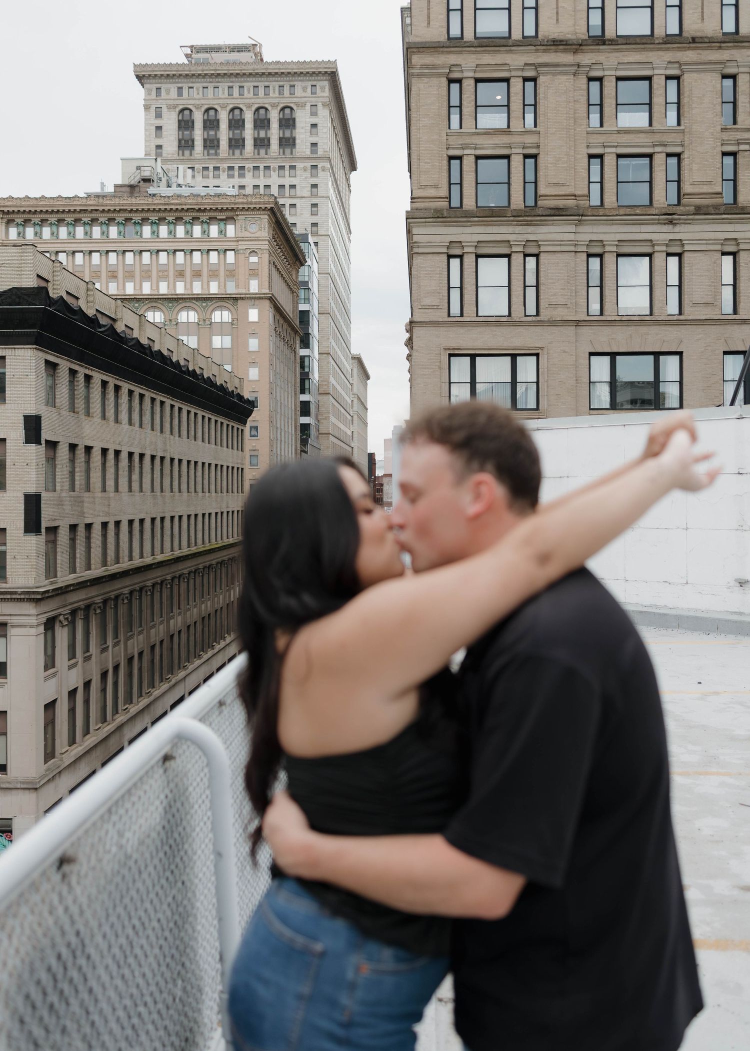 A romantic moment shared between a couple embracing on a rooftop with city buildings and architecture in the background.