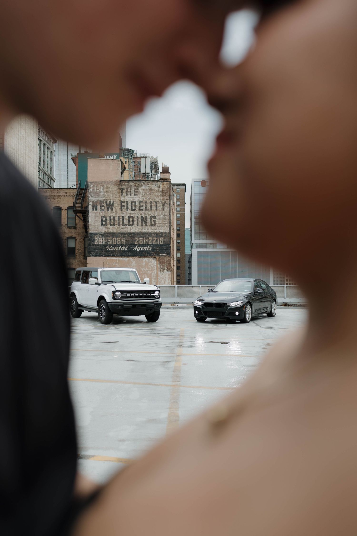 A blurred foreground frames a parking lot view of the New Fidelity Building with two parked cars in front.