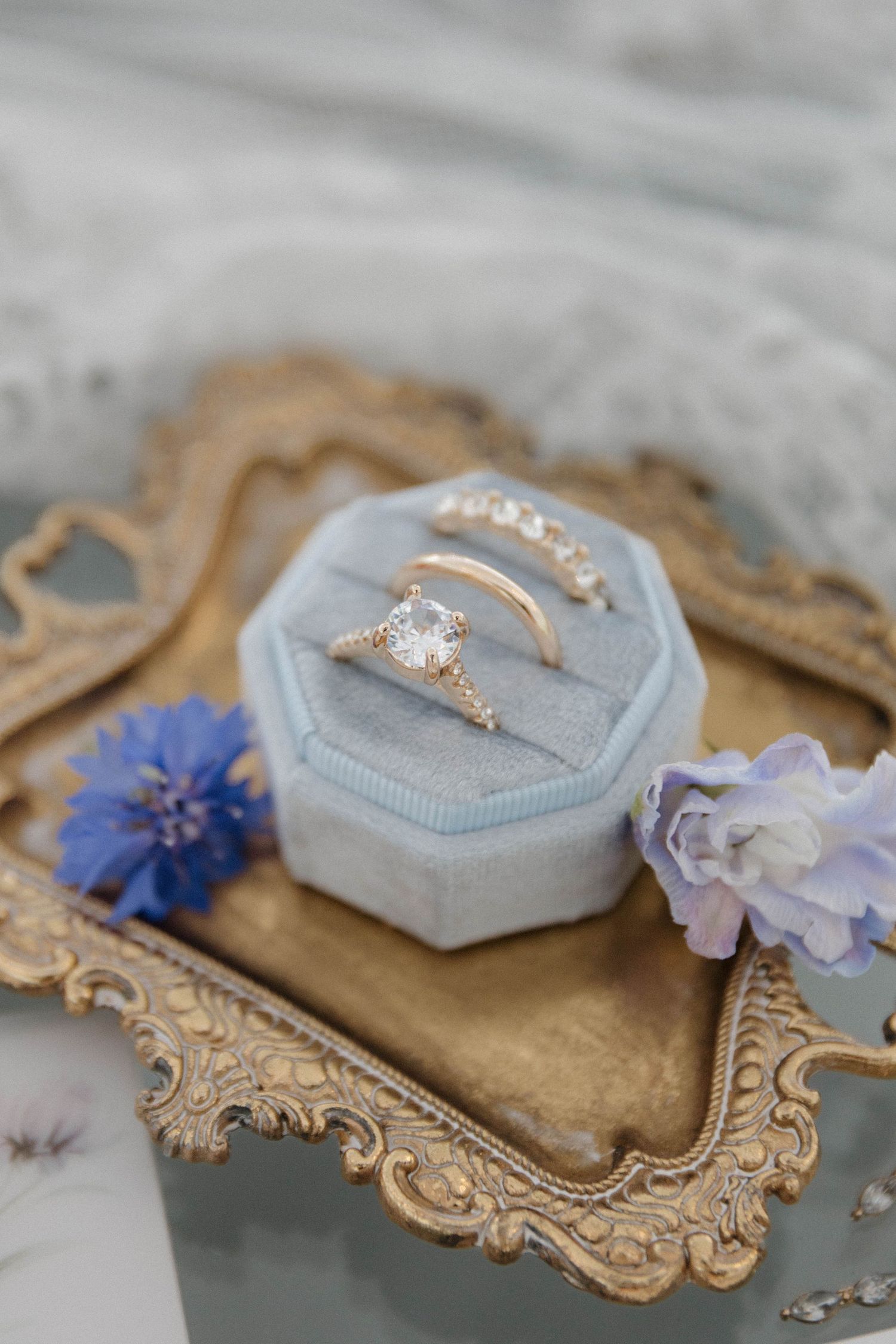 A velvet ring box with a diamond engagement ring displayed on an ornate gold frame with delicate purple flowers.