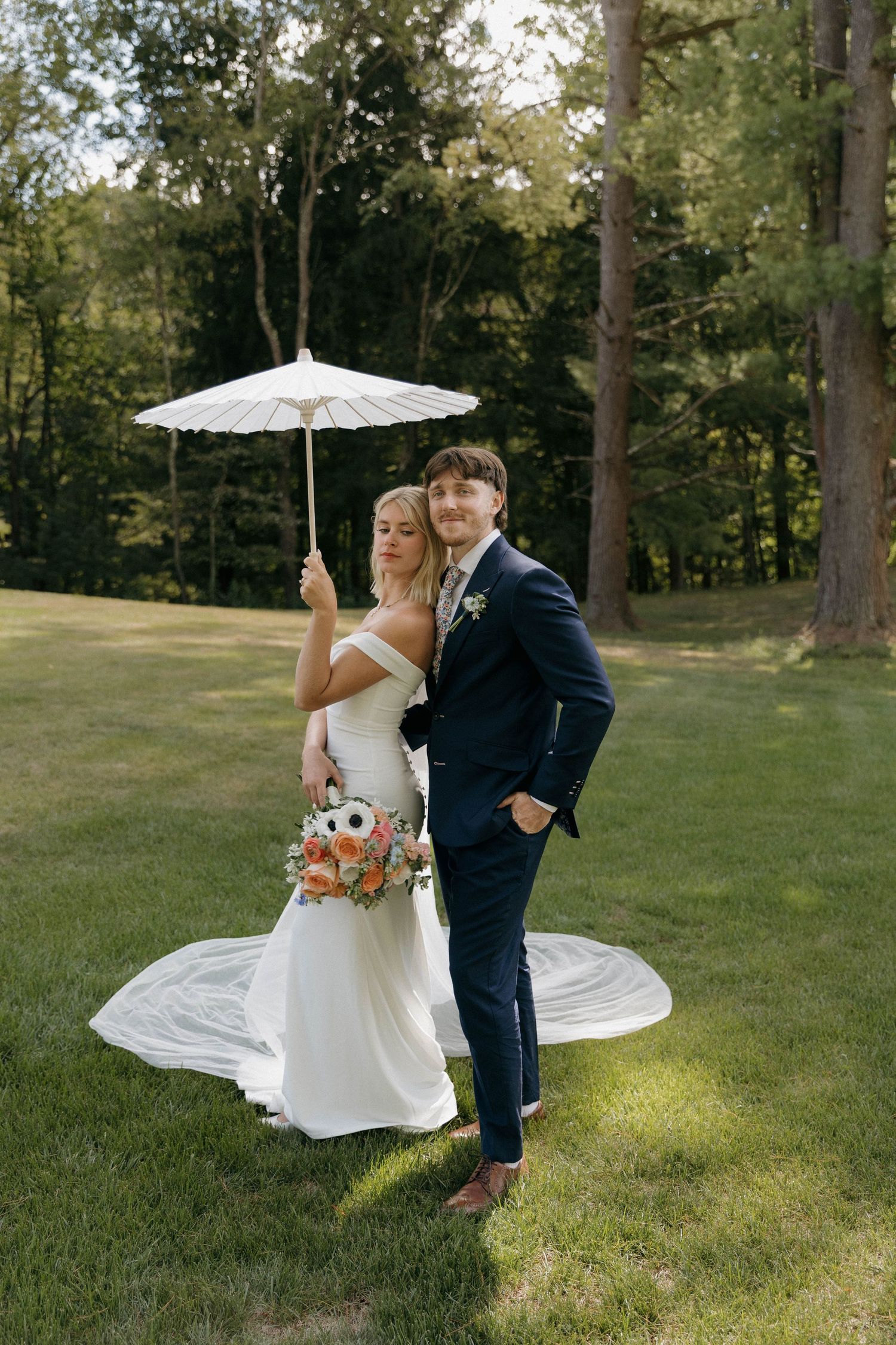 A wedding couple poses outdoors on a lawn with a white parasol and floral bouquet against a wooded backdrop.