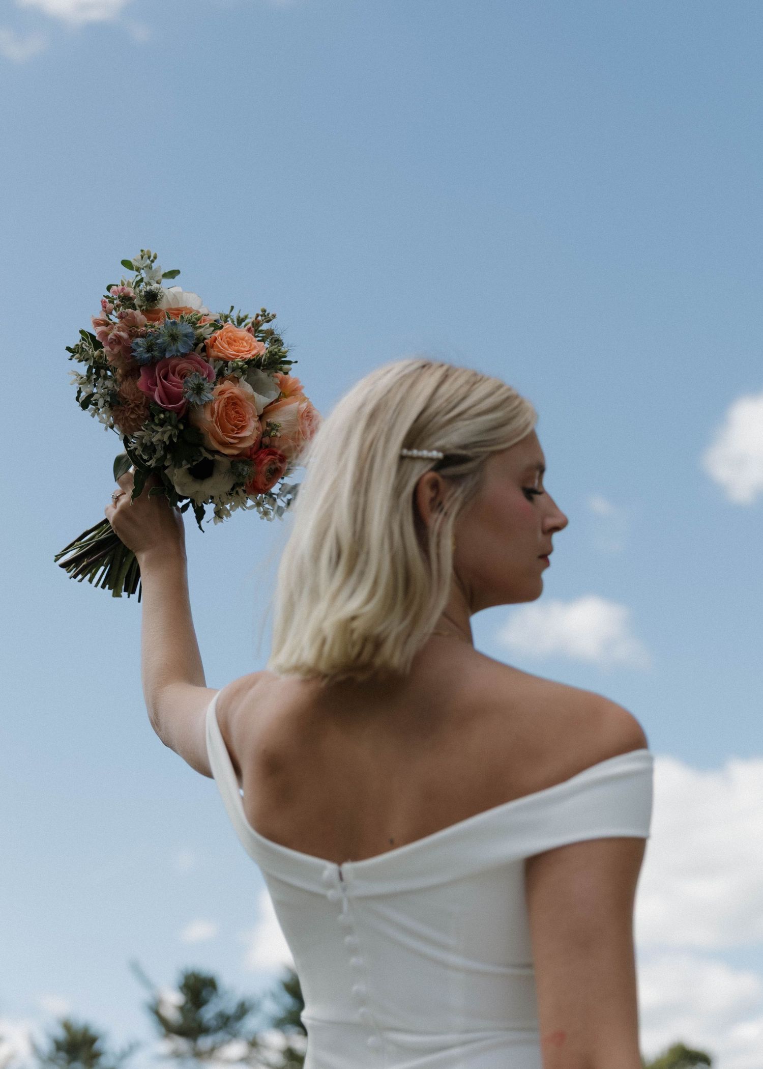 Person wearing an off-shoulder white wedding gown poses with a floral bouquet against a bright blue sky.