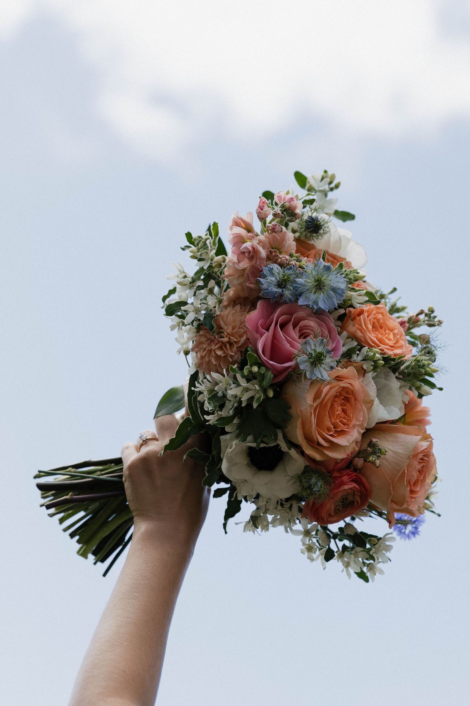 Colorful bridal bouquet held up against blue sky with peach roses and delicate wildflowers.