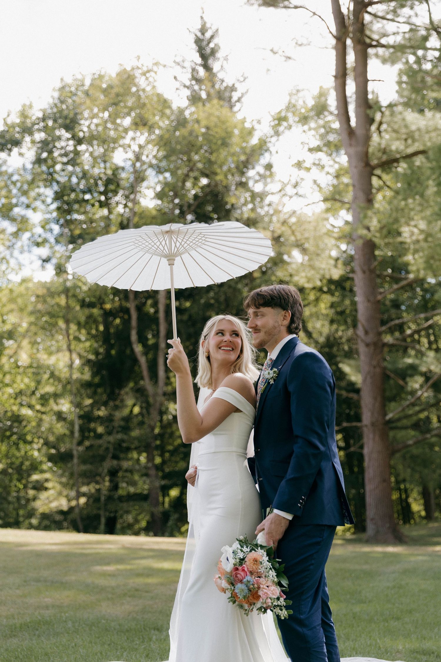 A couple shares joyful moments under a white parasol in a sunlit outdoor wedding setting.