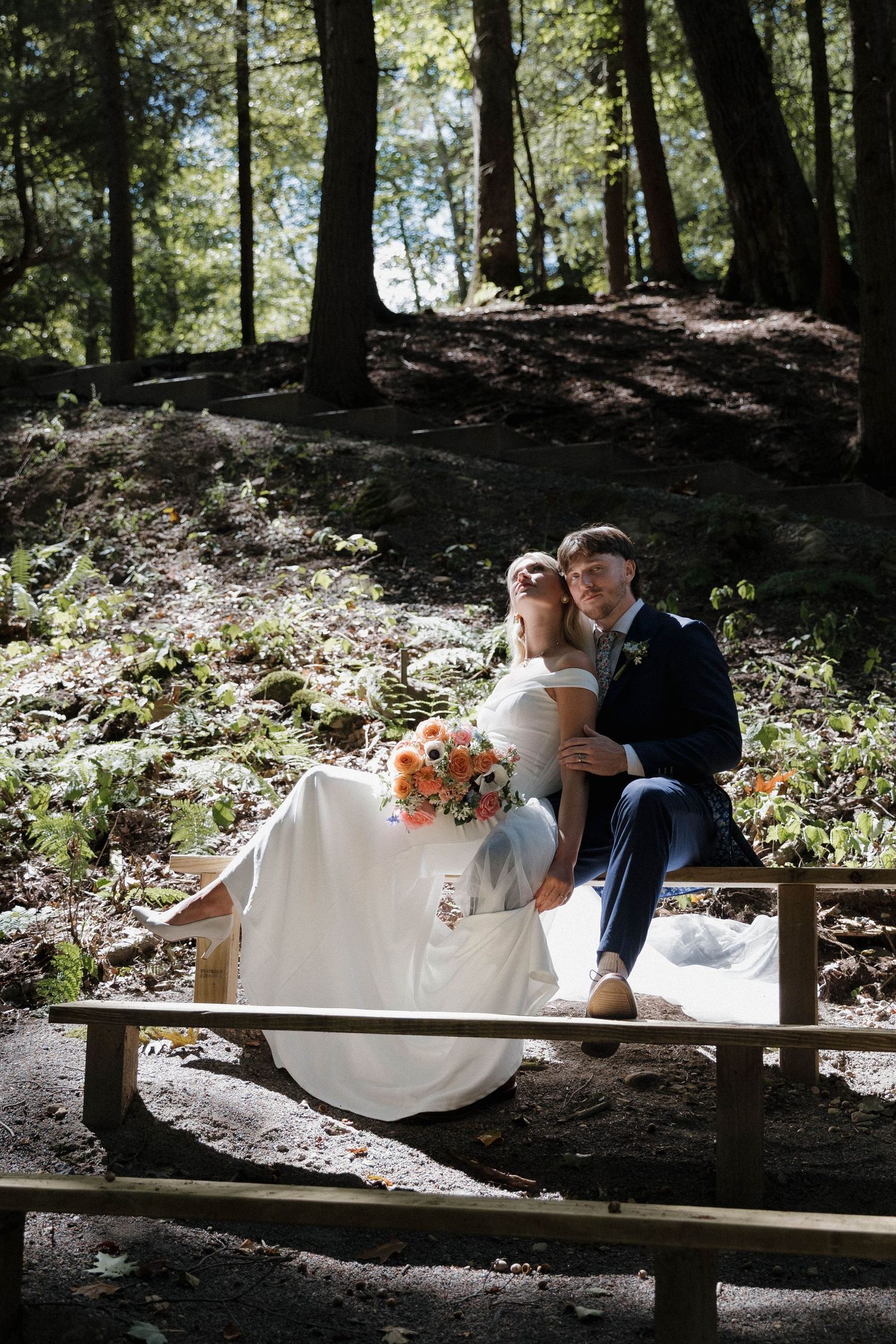 A couple shares tender moments on a wooden bench in a serene forest setting with floral decorations.