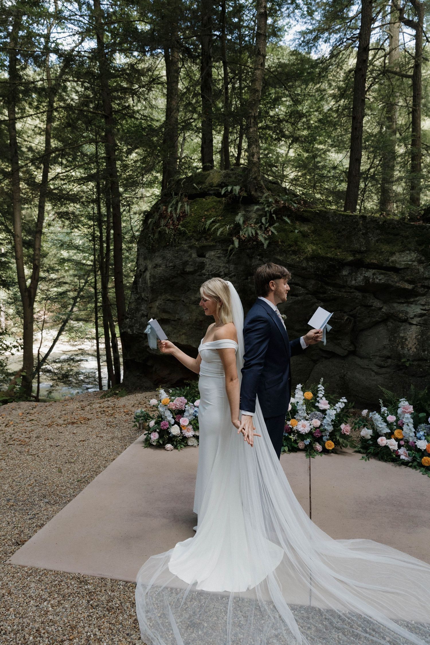 A wedding couple shares an intimate moment in the woods beside a floral arch and white parasol.