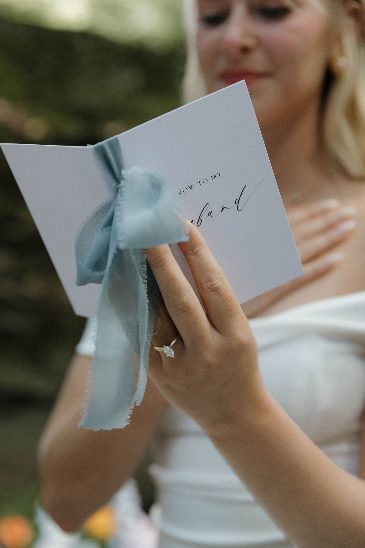 Hands gently hold a light blue letter envelope tied with ribbon during a wedding ceremony outdoors.