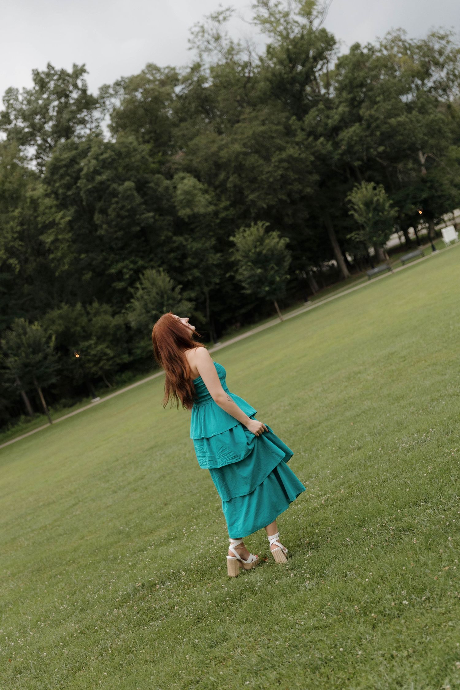 Figure in a flowing turquoise dress twirls on a grassy hillside with trees in the background.
