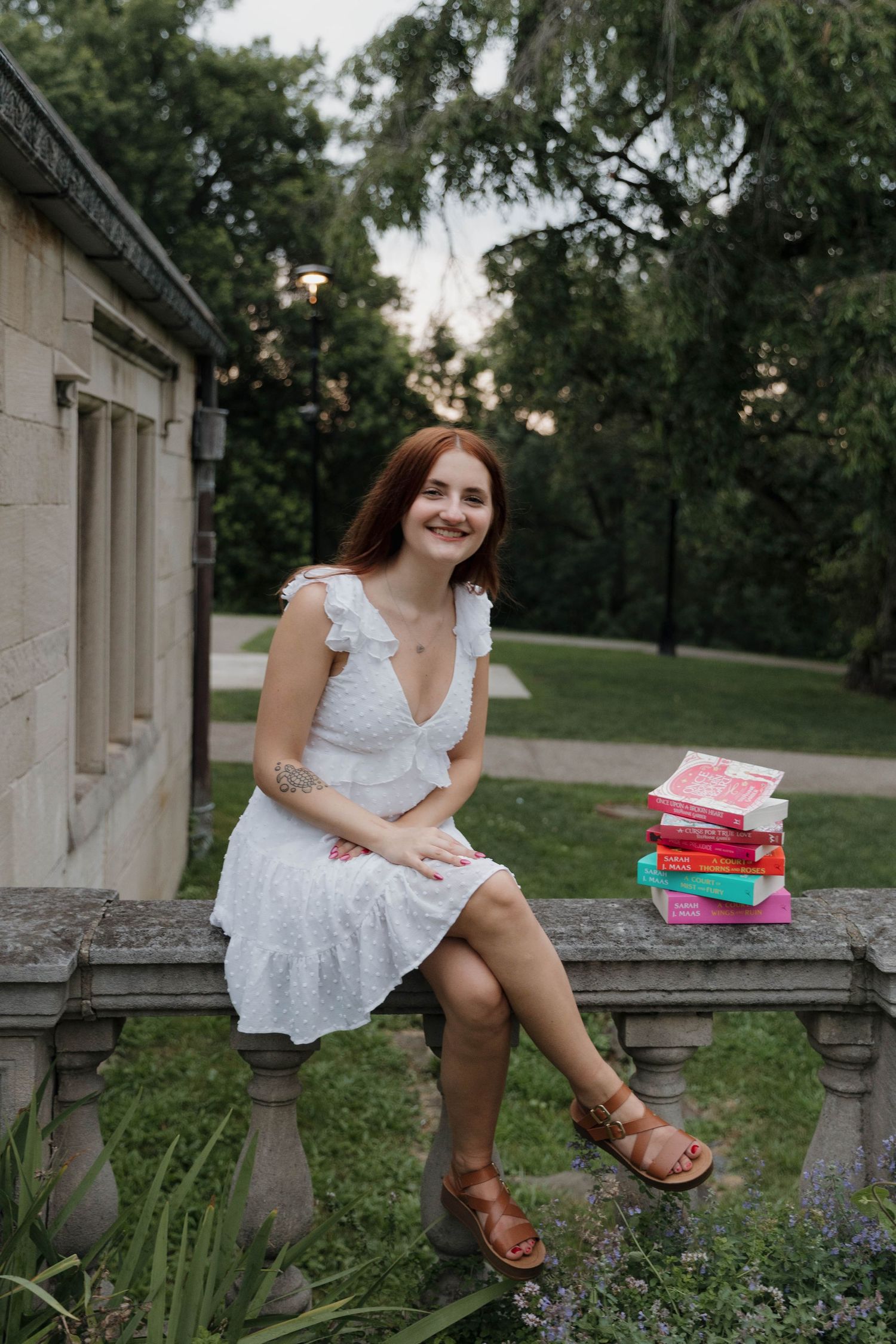 A white lace dress and brown sandals complement a stack of colorful books on a stone ledge in a garden setting.