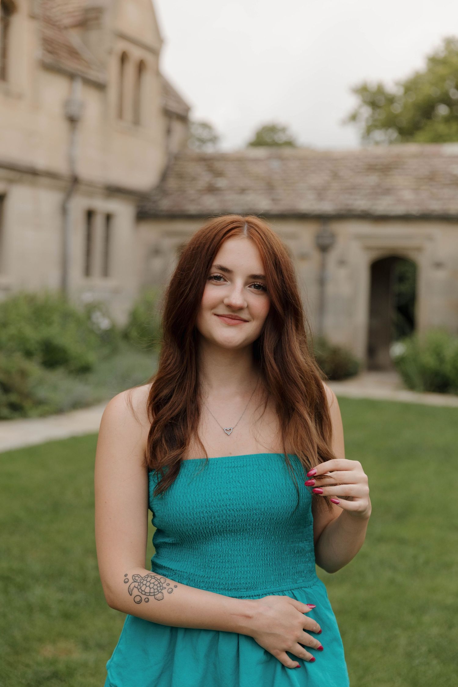 A person poses on a grassy lawn wearing a teal summer dress beside stone buildings in natural lighting.