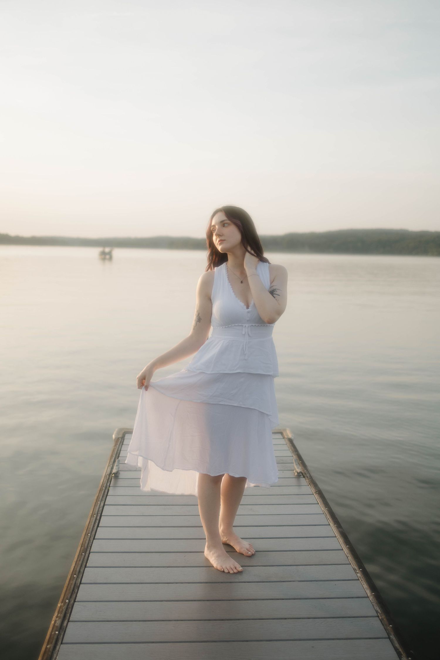 A barefoot person in a flowing white dress stands on a wooden dock overlooking a serene lake at sunset.