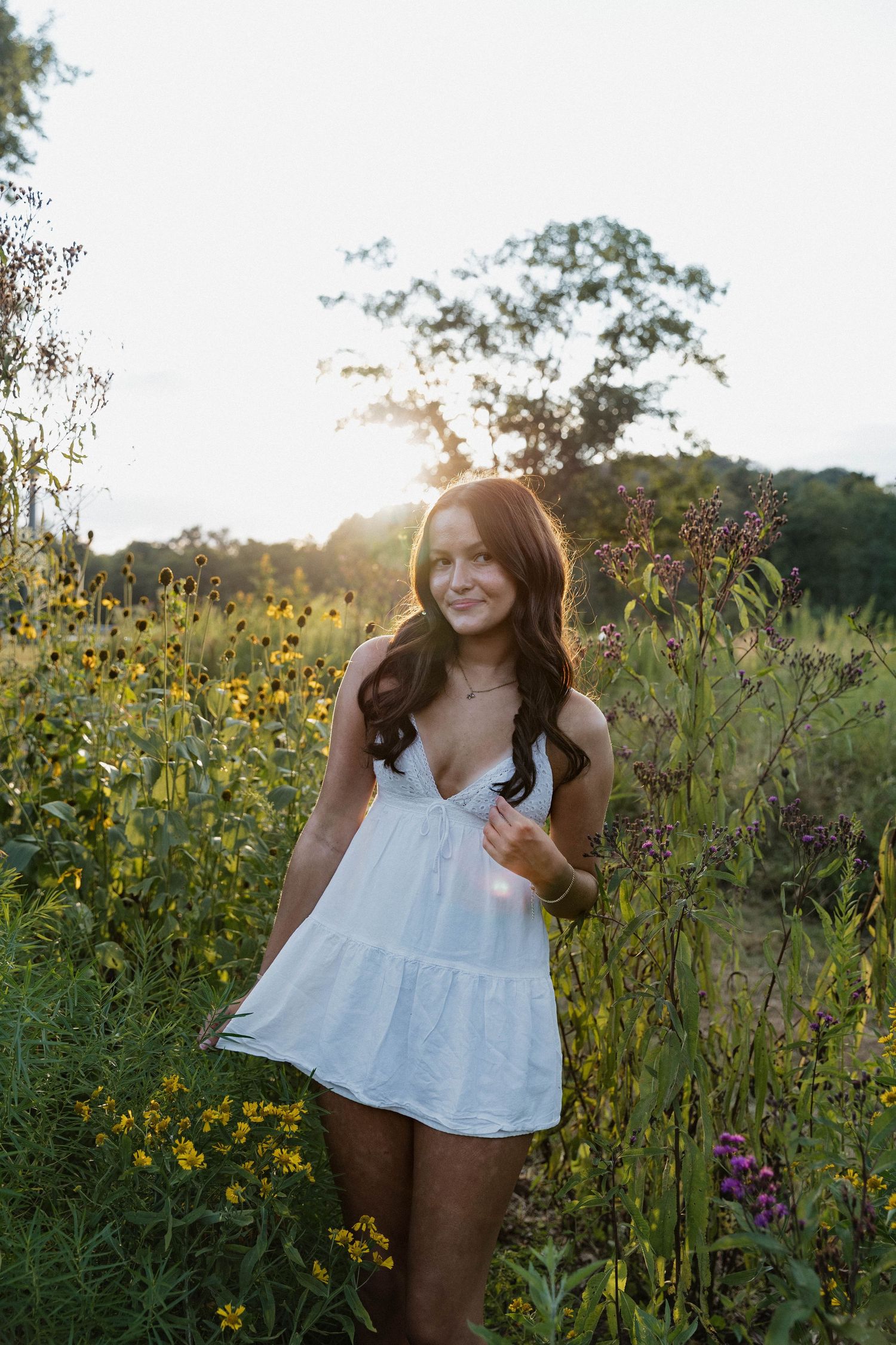 A white summer dress stands out against a golden sunset in a wildflower field filled with yellow blooms and tall grasses.