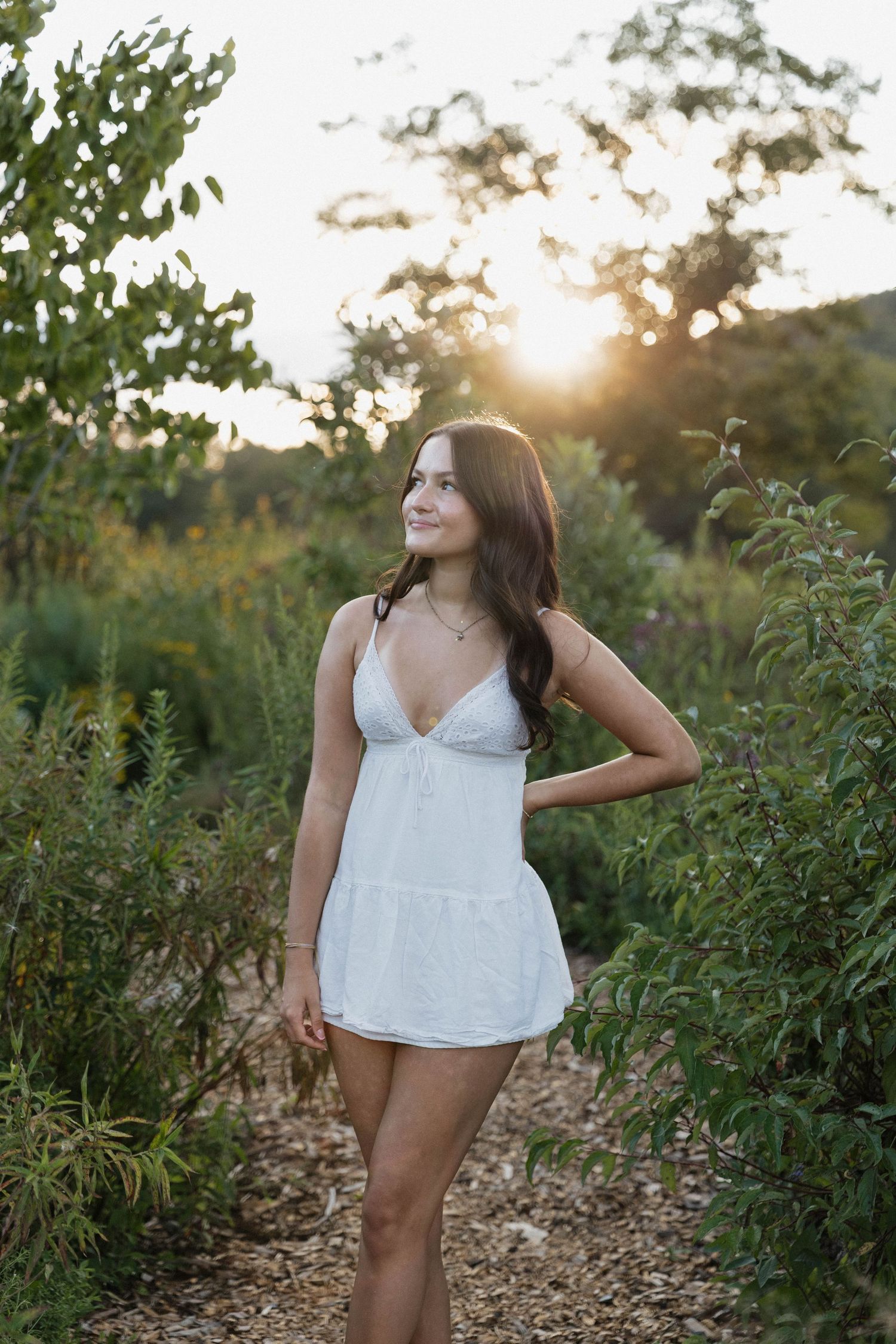Woman in white top poses in a sunlit field with tall wildflowers and greenery at golden hour.