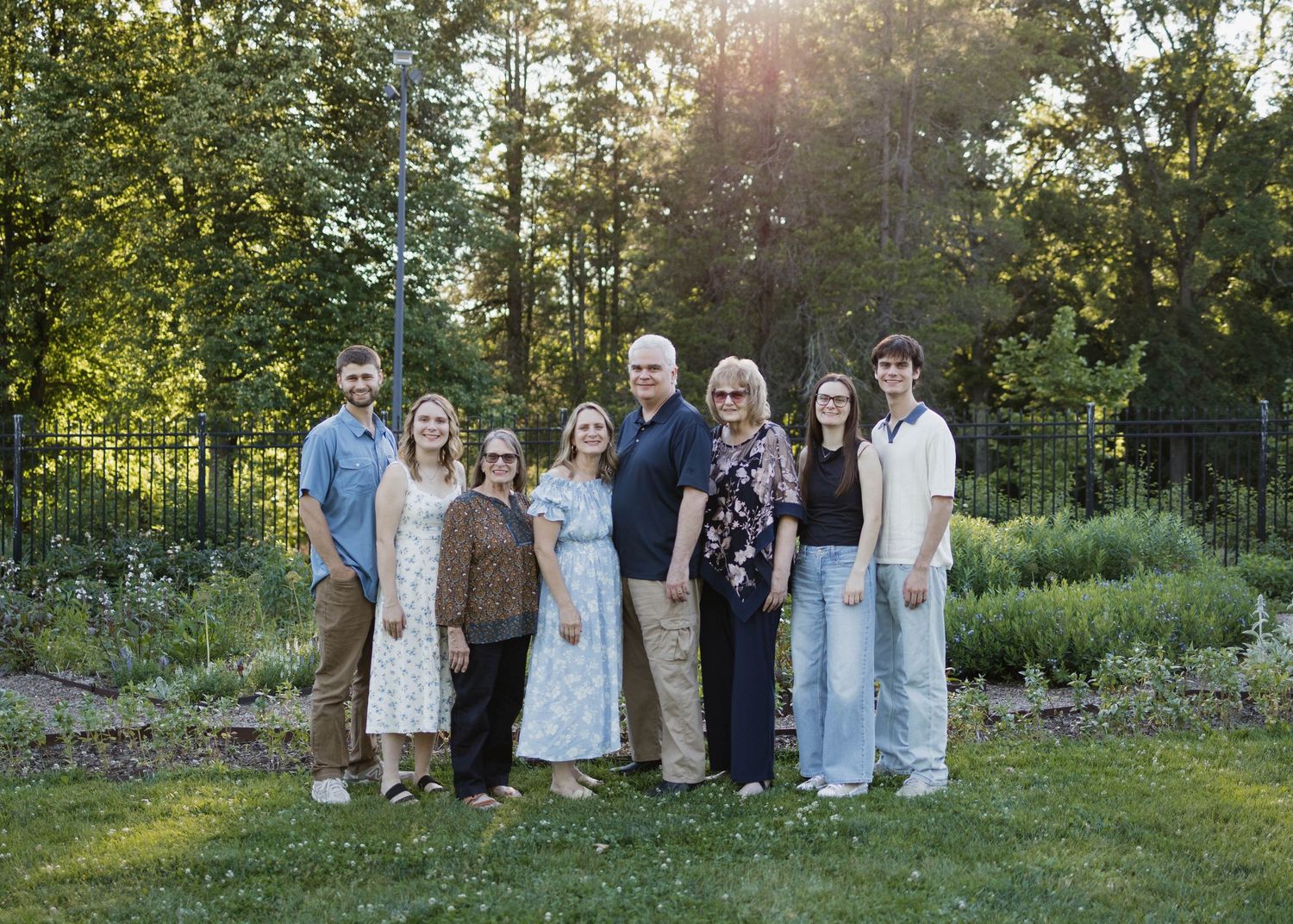A family of eight poses together outdoors on a green lawn surrounded by trees and garden beds during golden hour.
