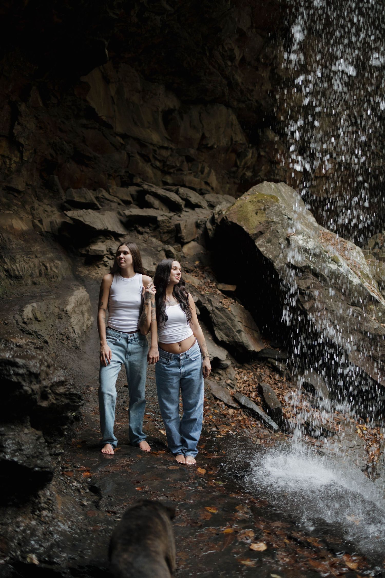 Two people in casual outfits explore a rocky cave area with a cascading waterfall in the background.