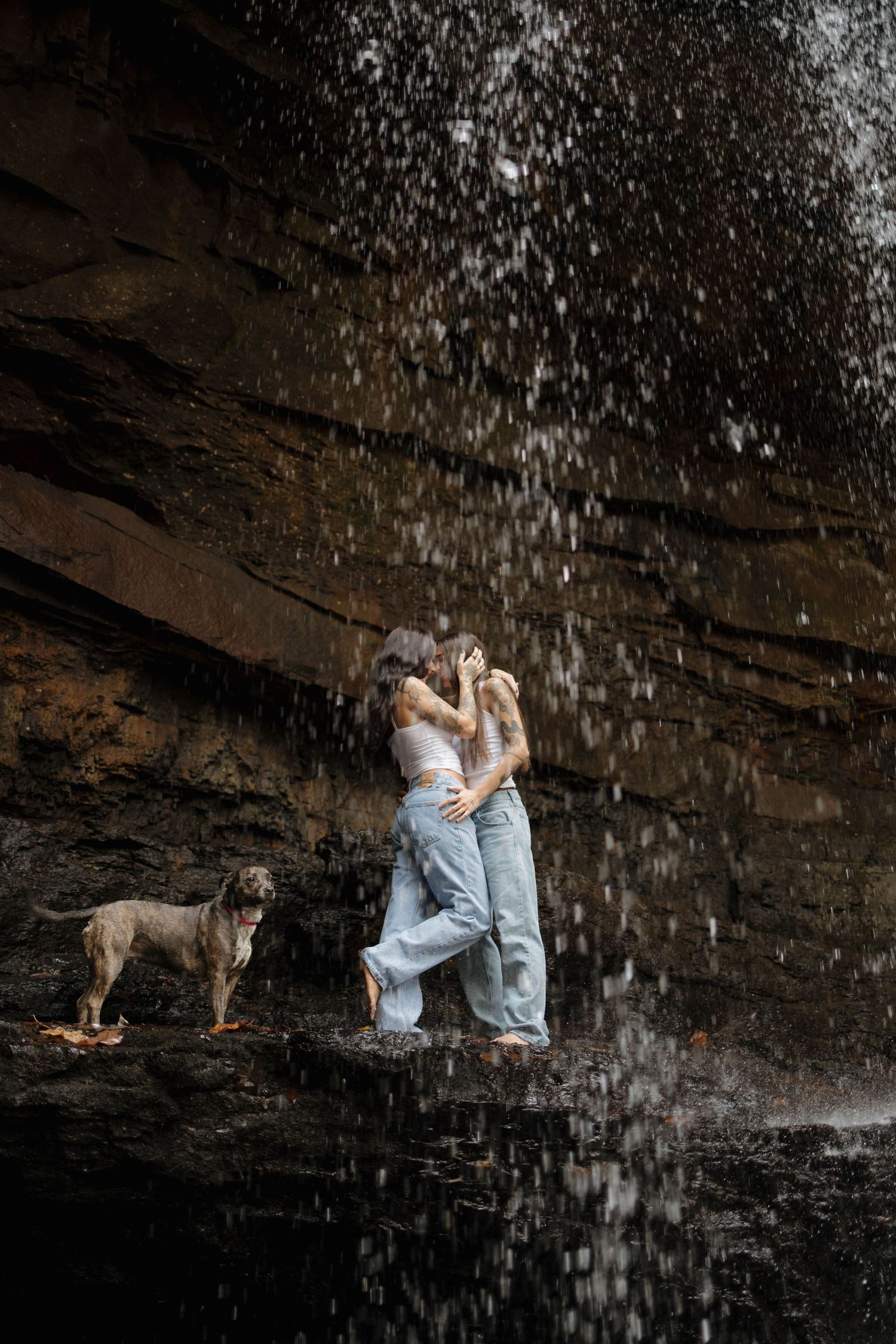 Two people sit beneath a rocky overhang with water cascading down in front of them while a dog watches nearby.